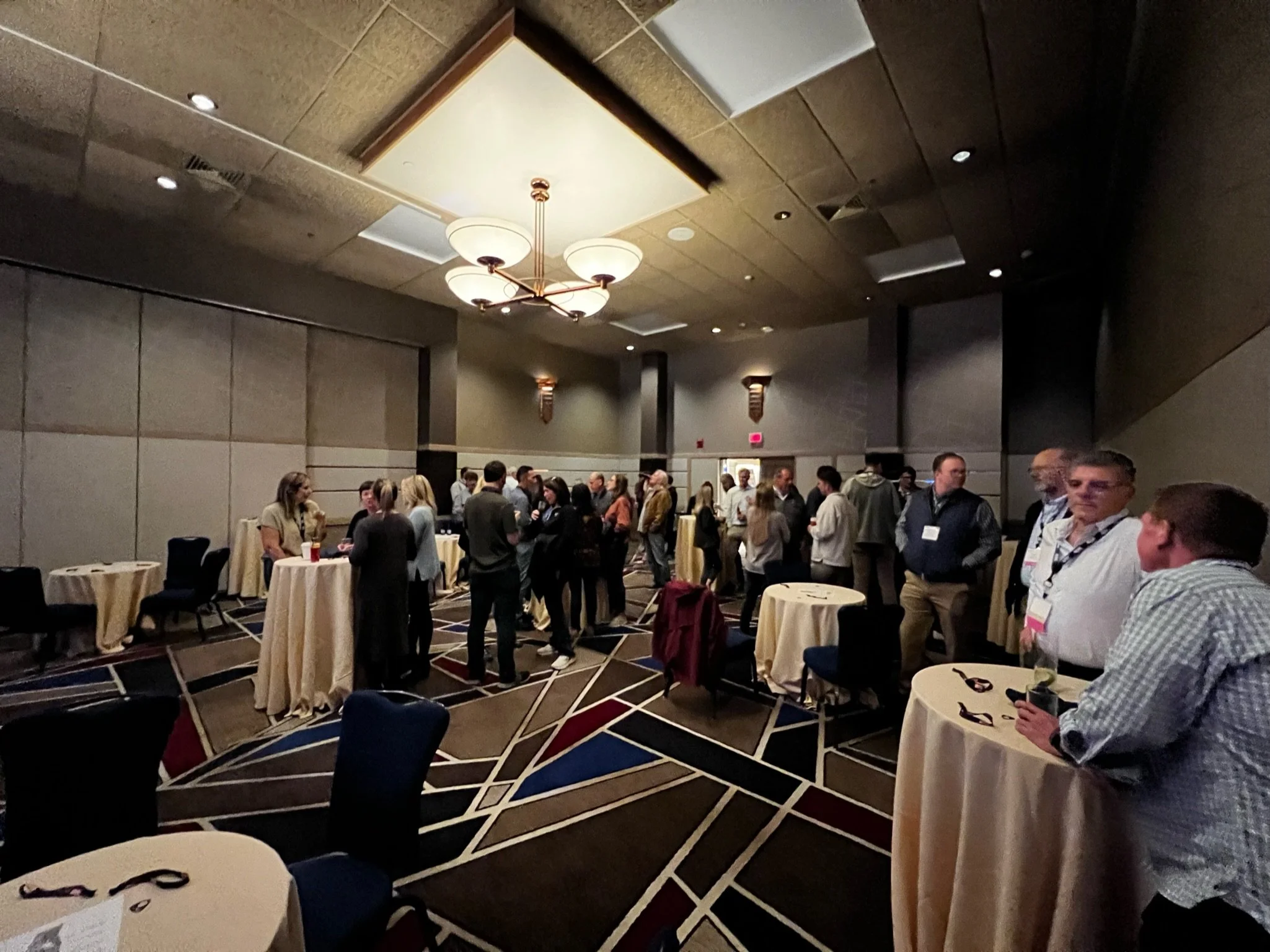 People standing and talking at cocktail tables in a large conference room with dim lighting and modern decor.