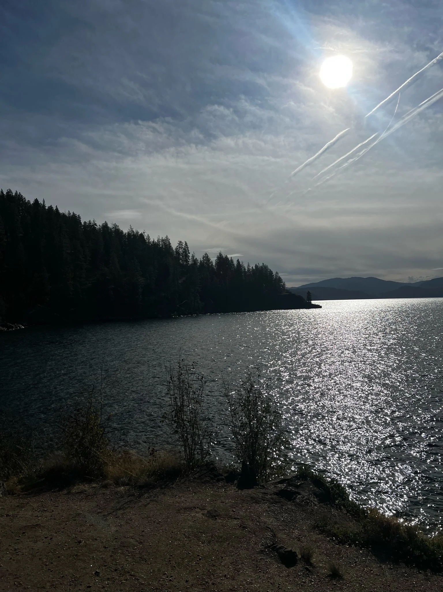 A scenic view of a lake with the sun shining in a partly cloudy sky, surrounded by forested hills and distant mountains.