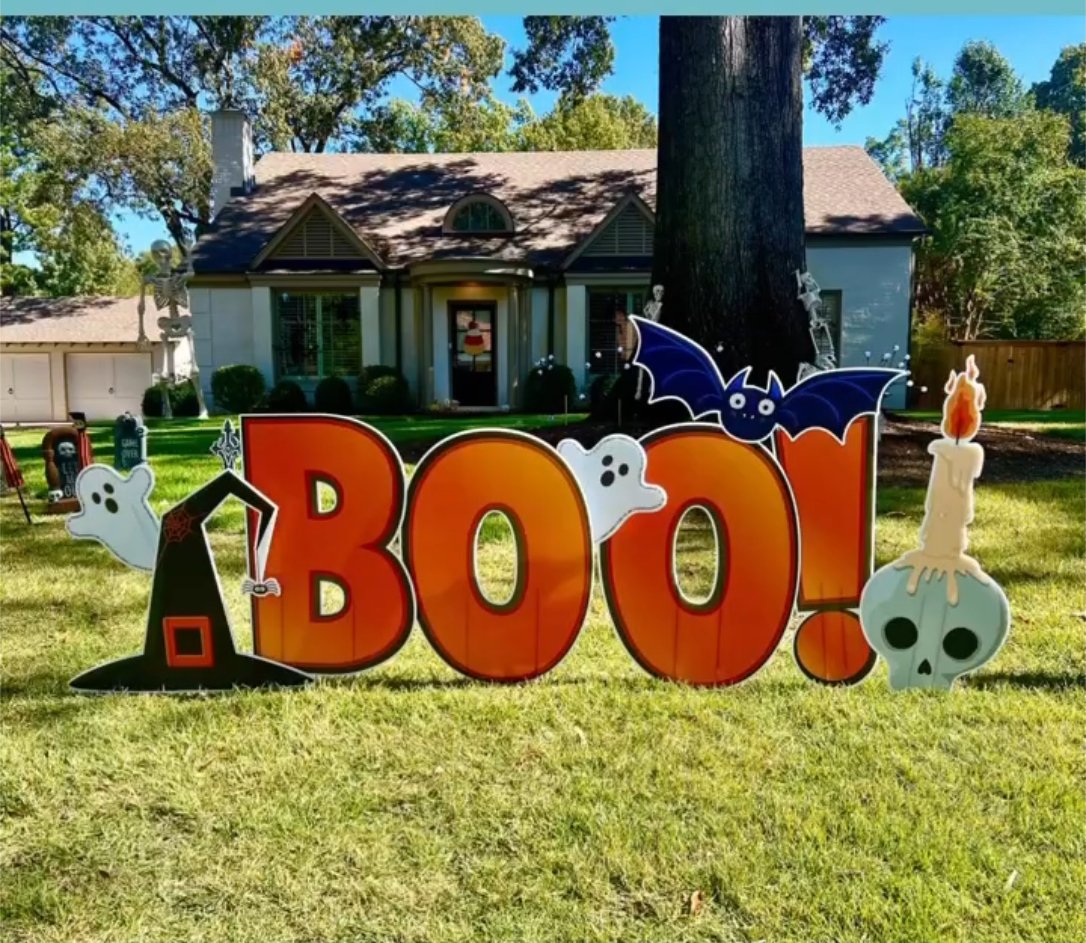 Halloween yard decoration with large orange 'BOO!' letters, a witch's hat, ghosts, bats, a skull with a candle, and skeletons around a large tree in front of a house.