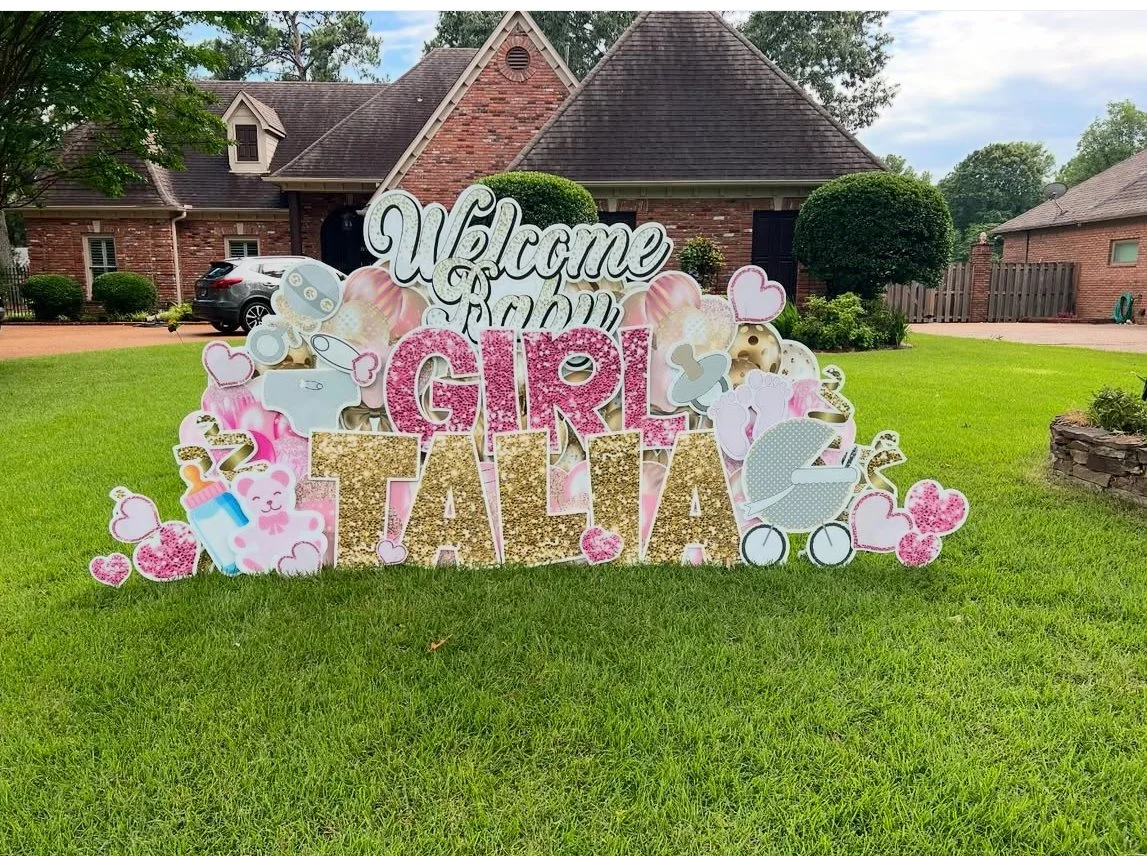 Decorative sign on a lawn welcoming a newborn girl named Talia, with baby-themed illustrations including hearts, a baby bottle, a teddy bear, and a baby stroller, in front of a brick house.