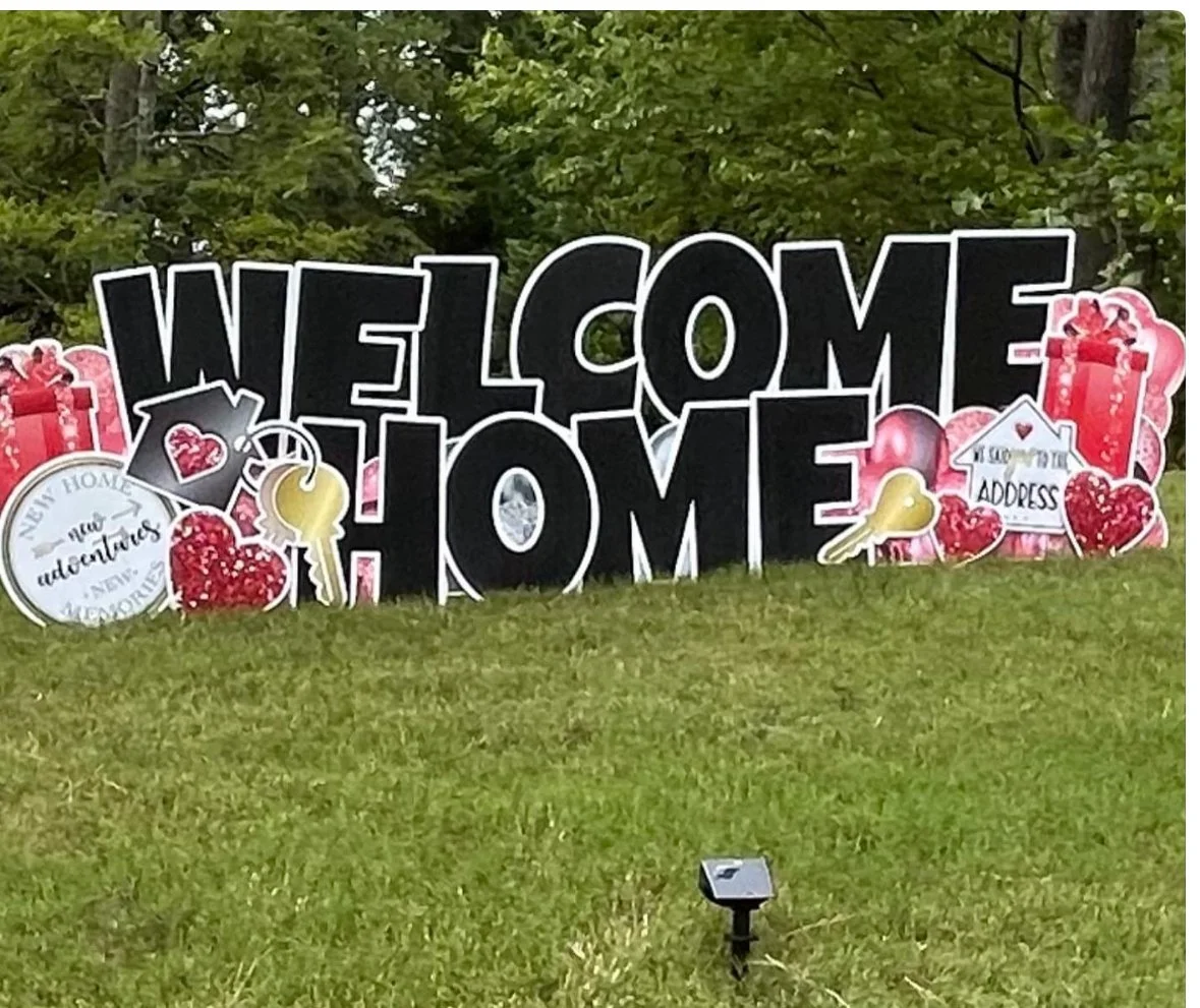 Decorative garden sign with large black letters reading "WELCOME HOME" surrounded by heart and key themed decorations on a lawn with trees in the background.