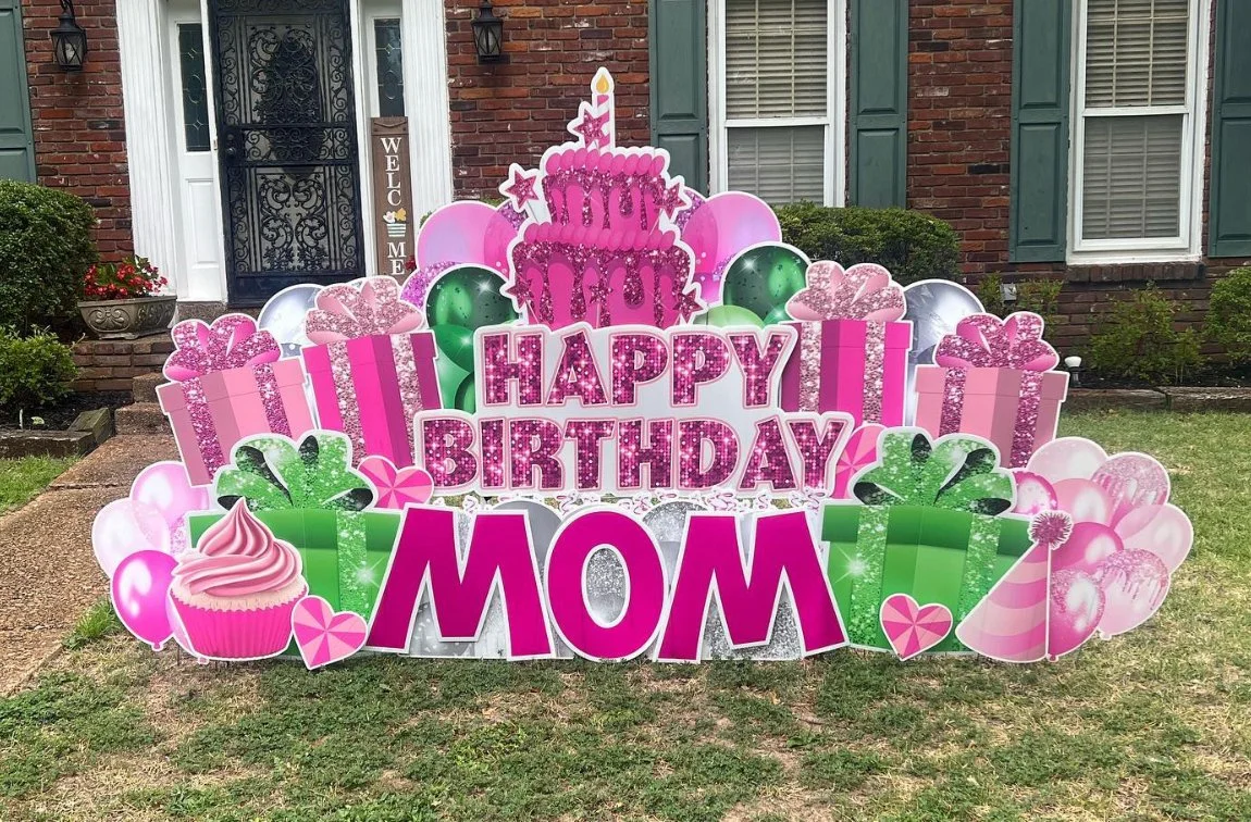 Colorful birthday display on a lawn with pink, green, and white decorations, including presents, balloons, a cupcake, and a large cake with a candle. The message reads "Happy Birthday Mom."
