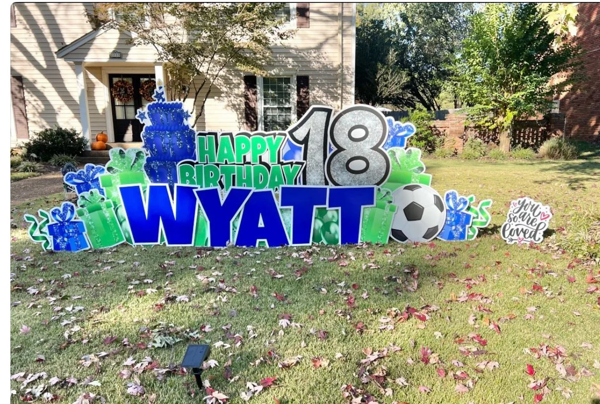 Decorative birthday yard sign celebrating an 18th birthday with a large blue cake, balloons, presents, a soccer ball, and signs with messages like "WYATT," "HAPPY BIRTHDAY," and "You are so loved" on a grassy lawn with fallen leaves.