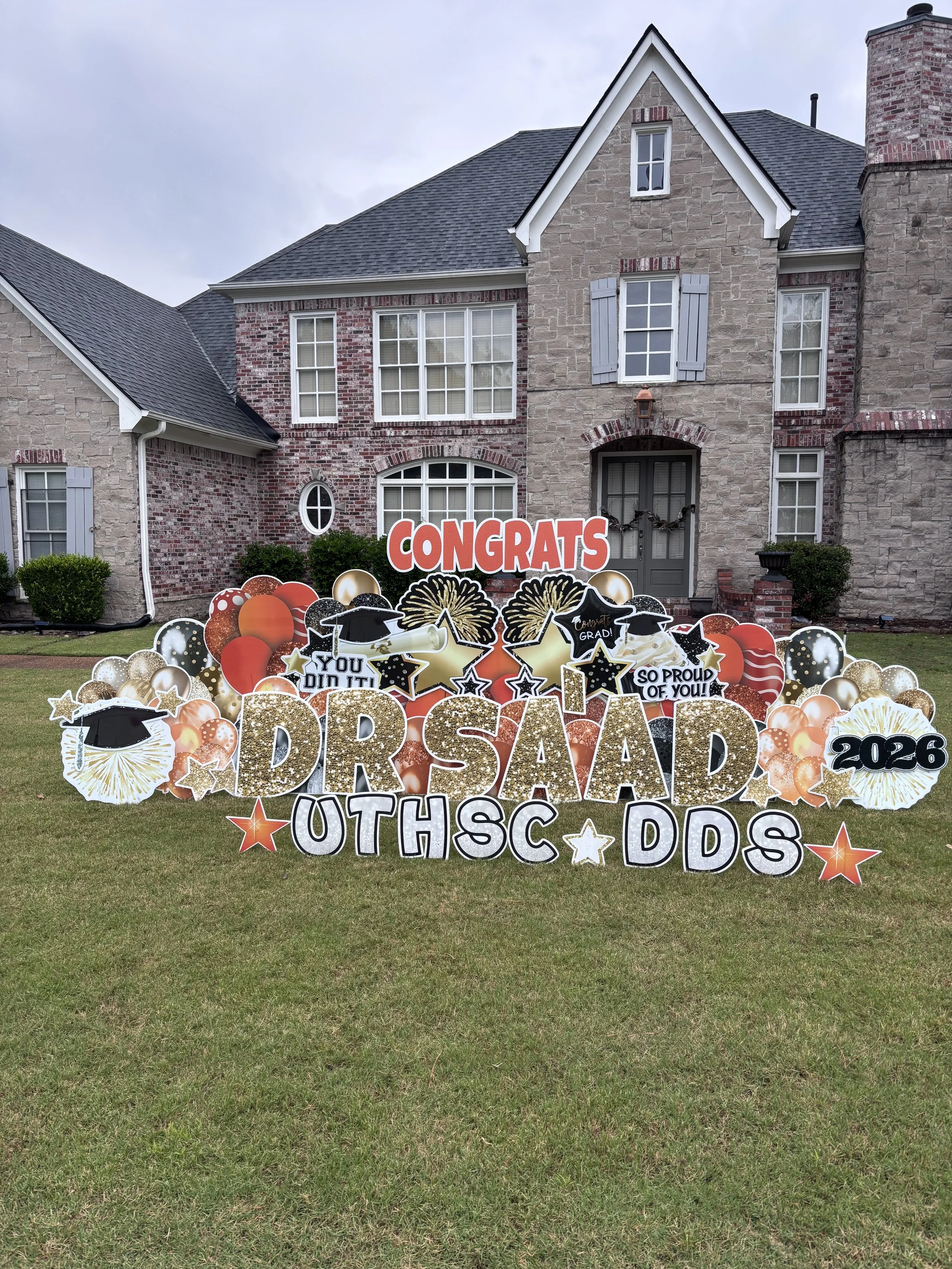 Front yard decorated to celebrate high school graduation, with a large sign saying 'Congrats Grad 2026' surrounded by stars, balloons, and graduation-themed decorations in front of a brick house.