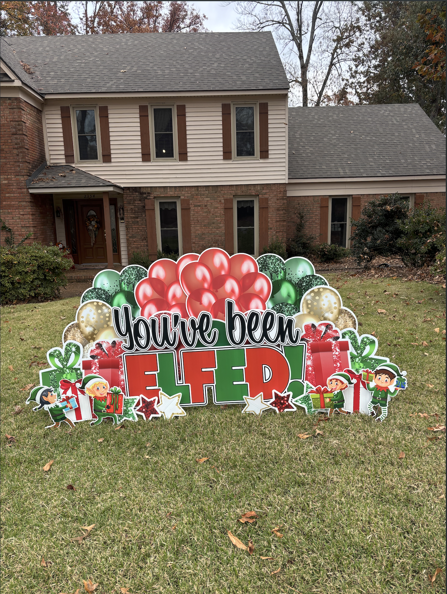 Festive holiday yard sign in front of a house with red brick and white siding, decorated with balloons, presents, elves, and holiday symbols, reading 'You've been Elfed!'