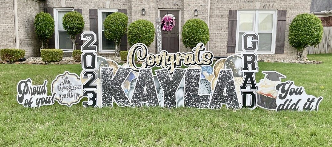 Graduation yard sign celebrating Kayla's graduation in 2023, with congratulatory messages, decorative elements, and a school-themed design, in front of a house with trimmed bushes and a pink wreath on the door.