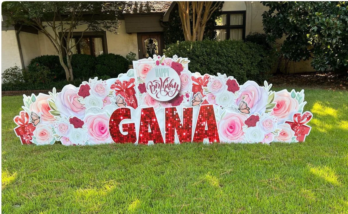 Garden display with pink and white roses, red roses, butterflies, gift boxes, and a sign that says "Happy Birthday." Large red letters spelling "GANA" are in front.