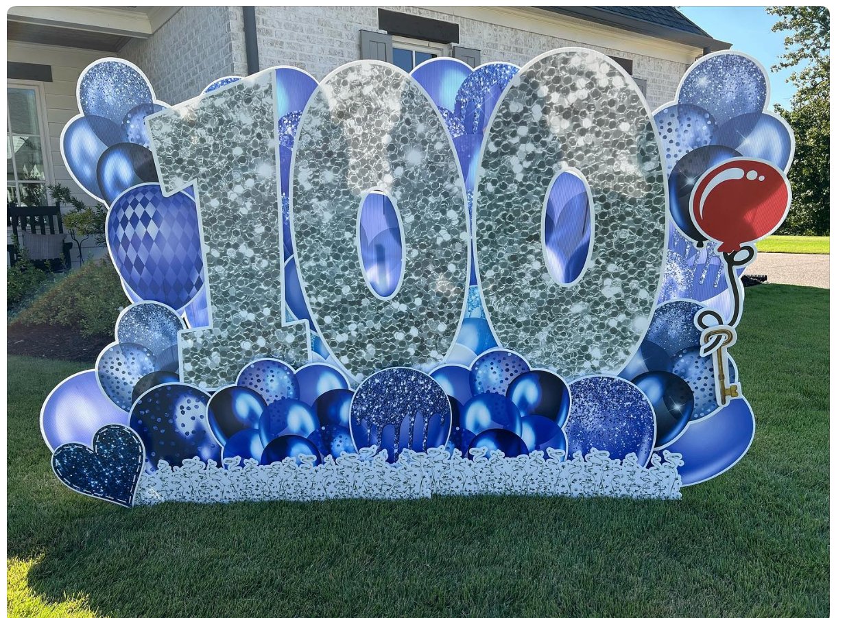 Decorative birthday display with large silver '100' numbers surrounded by blue and silver balloons and a red balloon with a white highlight, set up outdoors on a lawn in front of a house.