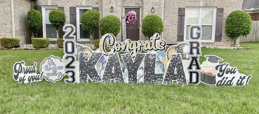 Graduation congratulation display on a lawn with large decorated cutouts reading 'Congrats Kayla,' '2023,' 'Grad,' and motivational phrases. The background shows a house with potted trees, windows, and a door with a wreath.
