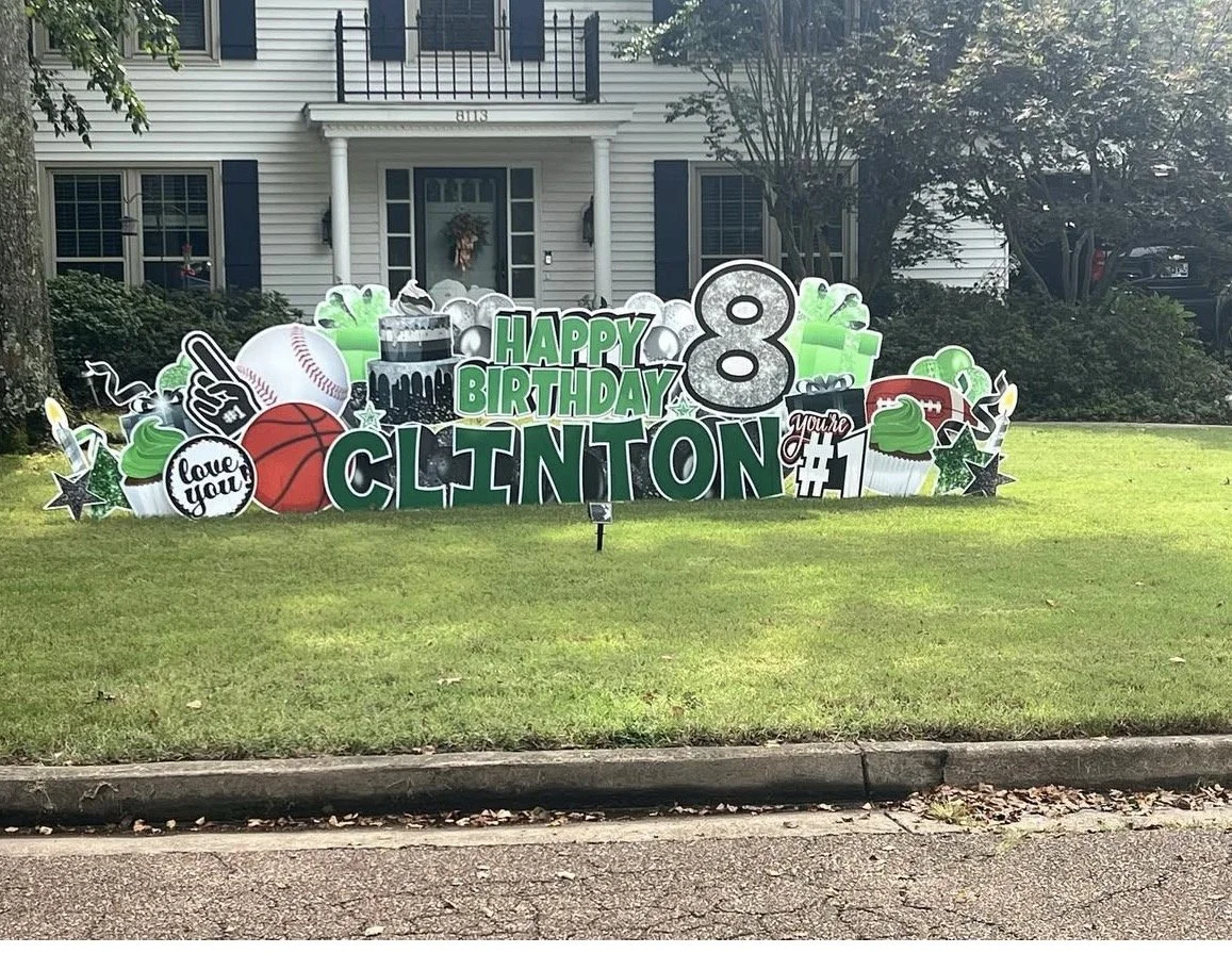 Large birthday yard decoration celebrating an 8th birthday for Clinton, themed with sports and love icons, including a baseball, basketball, football, cupcake, sports equipment, and a cake, placed in front of a house with a green lawn.