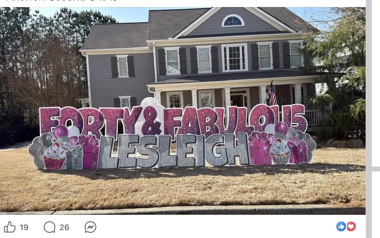 Large decorative yard sign celebrating Leslie's birthday with words "Fabulous & Forties Leslie" in pink and silver letters, surrounded by balloons and cupcake images, in front of a gray two-story house.