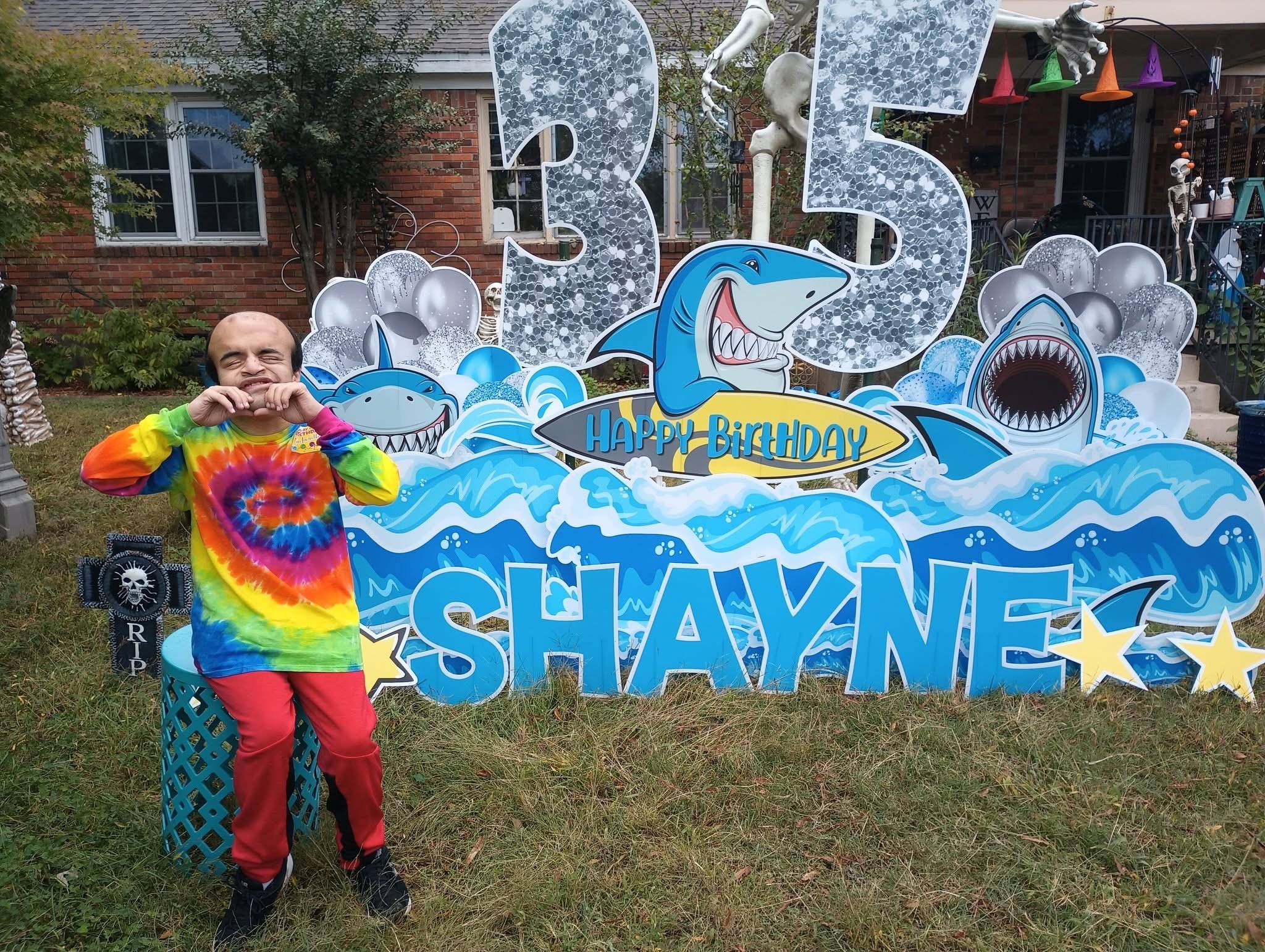 A young boy with curly hair, wearing a colorful tie-dye shirt and red pants, standing in front of a birthday party decoration with a shark theme. The decoration features large numbers '3' and '5' with shark graphics, water, and balloons, and a sign t