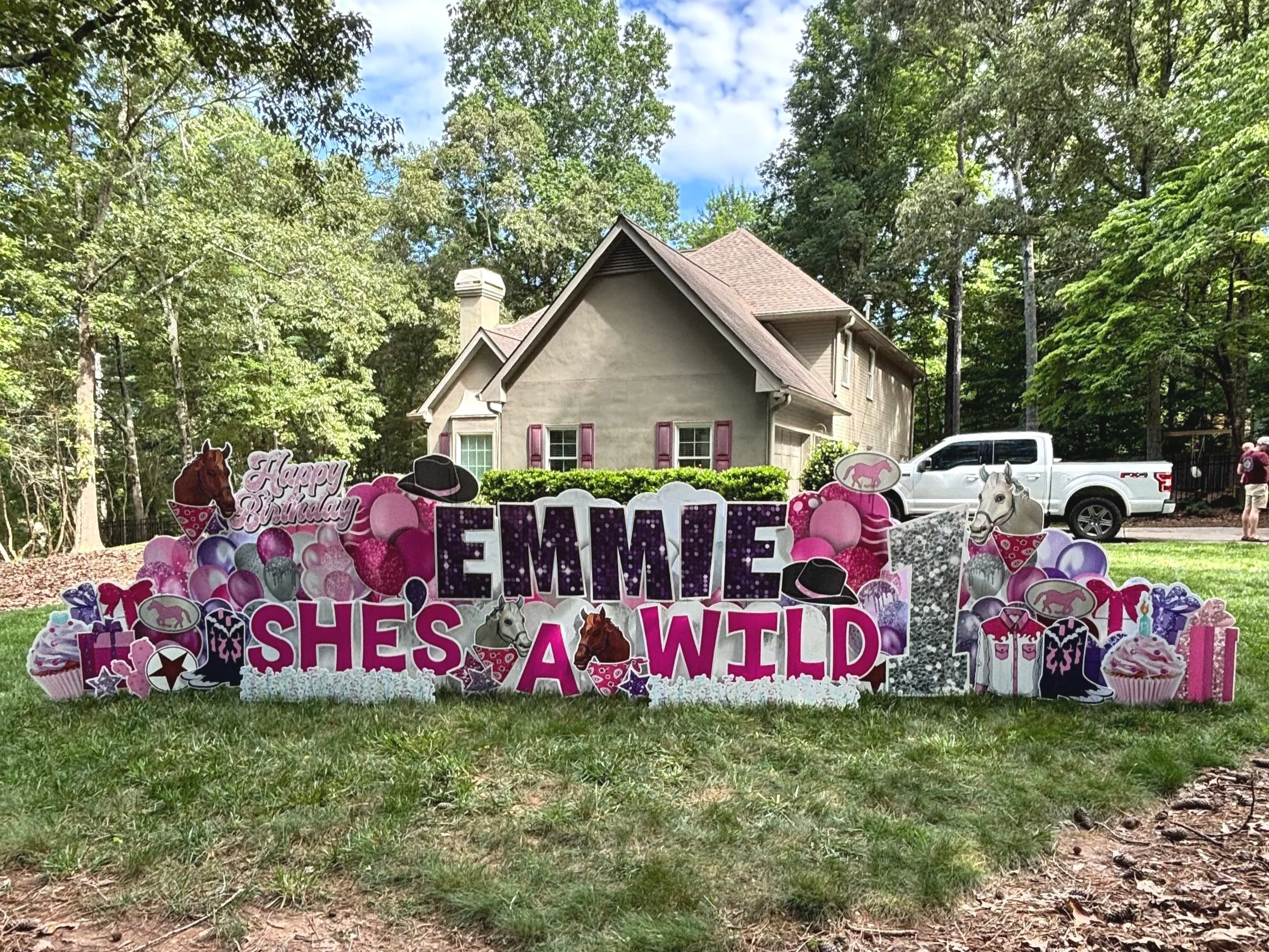 Decorations for a birthday party with a pink theme featuring cutouts of horses, cupcakes, hot pink balloons, and words like 'Happy Birthday', 'Emmie', 'Sweet', and 'Shes & Wleft' in a yard in front of a house surrounded by trees.