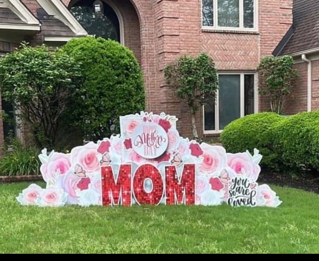 Decorative arrangement on a lawn with large red letters spelling 'MOM', surrounded by pink and white flowers, with signs reading 'Mother's Day' and 'You are so loved', in front of a brick house.
