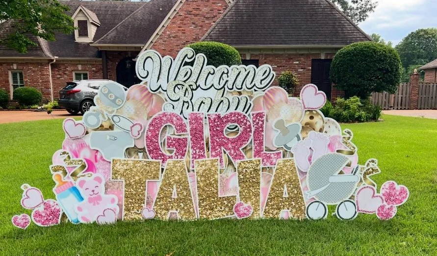 Decorative sign celebrating a baby girl named Tatiana, featuring the words "Welcome Baby Girl Tatiana" with baby-themed designs like hearts, a baby bottle, a stroller, a teddy bear, and cupcake illustrations, set on a grassy lawn with houses in the b