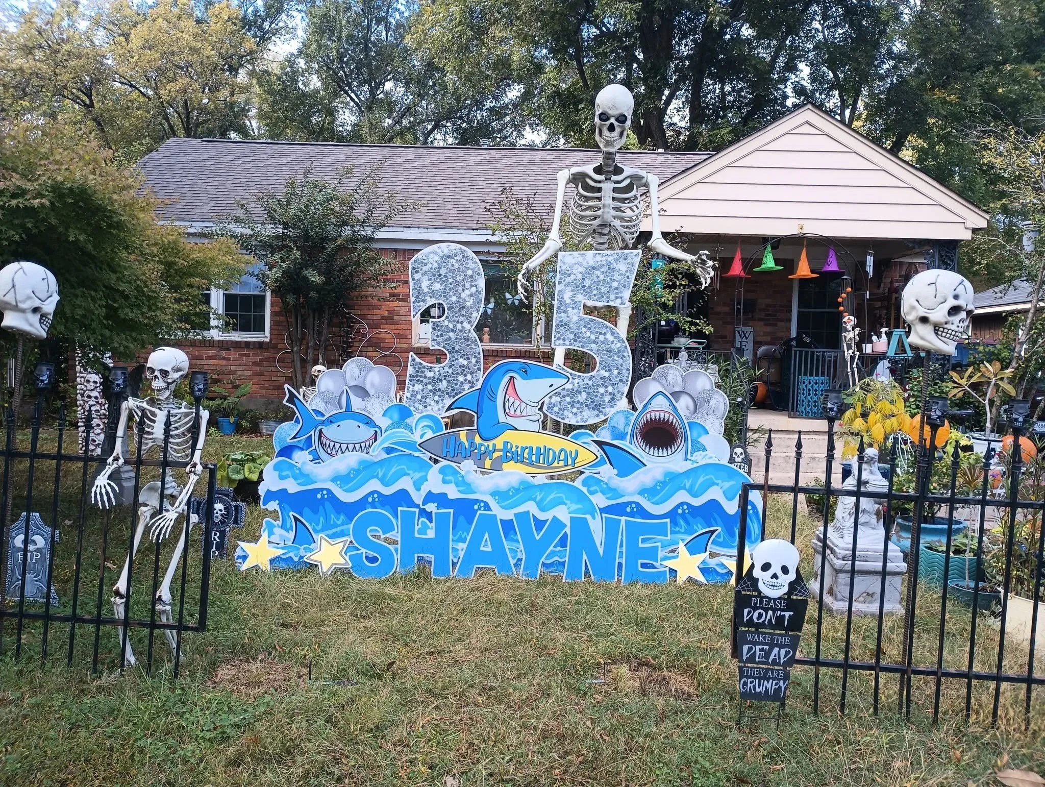 Decorative Halloween scarecrow display in a front yard with skeletons, sharks, and colorful signs, including large letters spelling 'SHANE' and number '3'. There are inflatable skulls, hanging neon hats, and a sign that reads 'Please don't wake the d