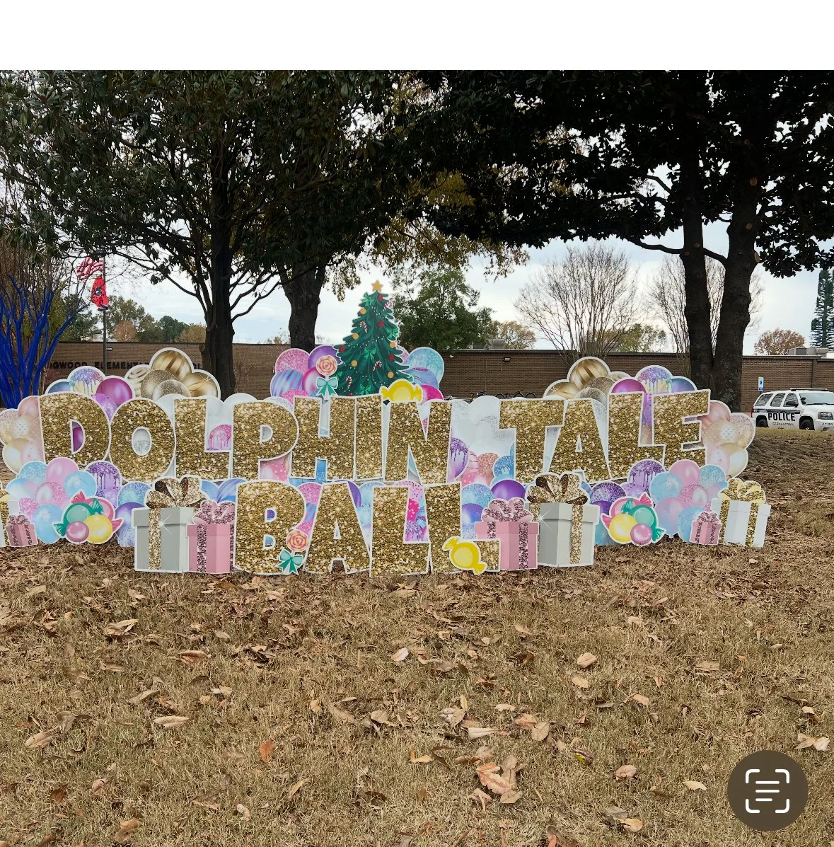 Decorative sign reading 'DOLPHIN TALE BALL' surrounded by balloons, gift boxes, and a Christmas tree cutout, set outdoors on grassy ground with trees and a brick building in the background.