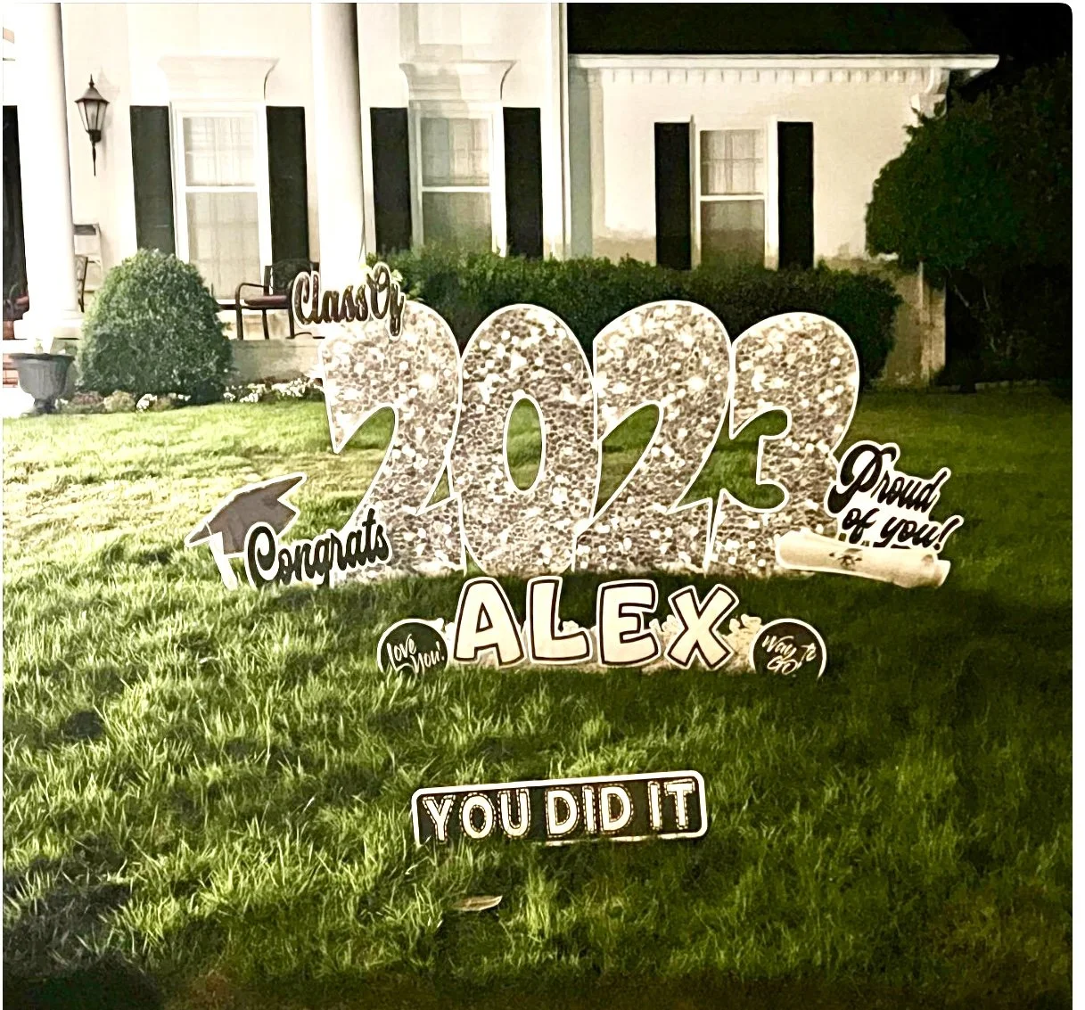 Decorative graduation yard sign celebrating a 2023 graduate named Alex, with congratulatory messages and encouragement, placed on a grassy lawn in front of a house at night.