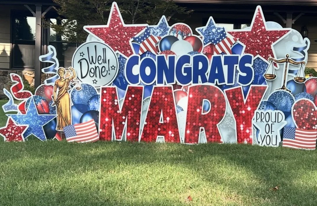 Decorative sign celebrating a graduation, featuring the words 'Congrats Mary,' with patriotic red, white, and blue colors, stars, American flags, a statue of justice, and the phrases 'Well Done!' and 'Proud of You.'