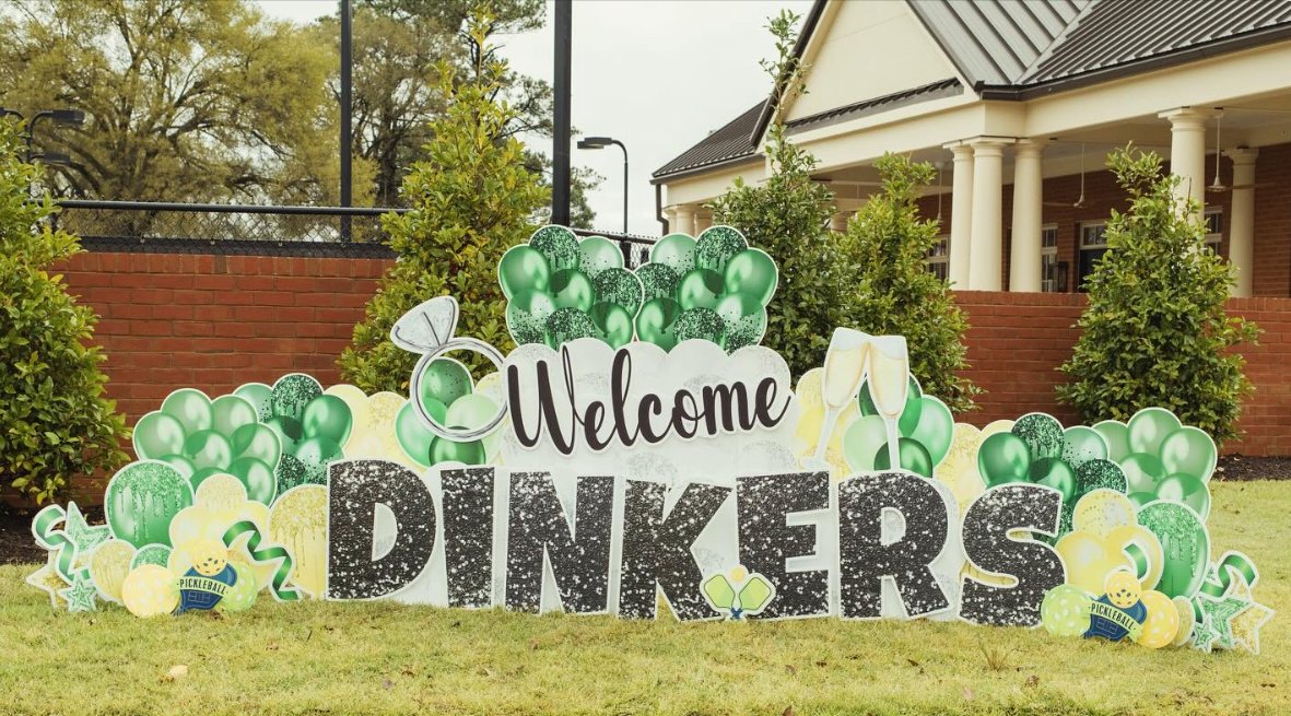 Decorative sign on grass reading 'Welcome DINKERS' with balloons, champagne glasses, and other festive decorations for a celebration.