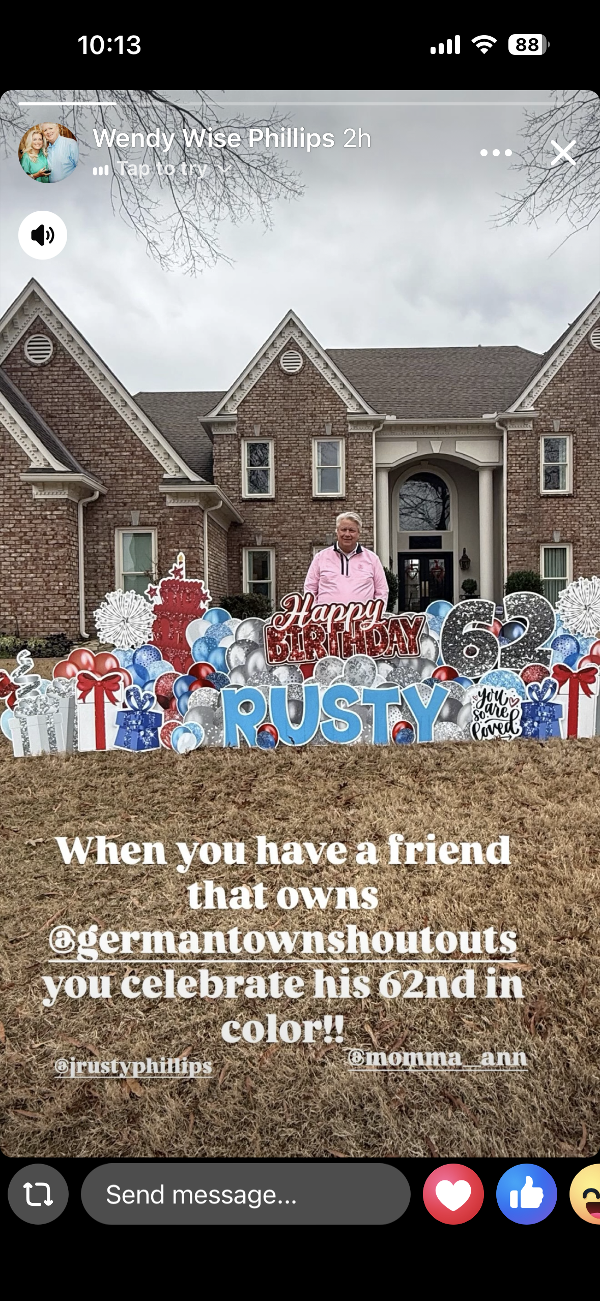 A man standing in front of a decorated lawn celebrating his 62nd birthday, with a house and bare trees in the background. Decorations include balloons, gift cutouts, a birthday cake cutout, and a sign reading 'Happy Birthday Rusty'.