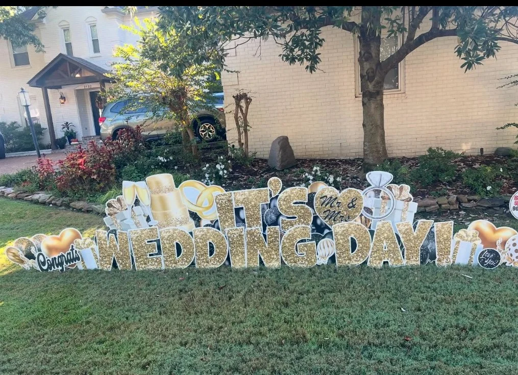 Outdoor wedding celebration display with large gold and black glitter signs reading 'Congratulations Mr. & Mrs. Wedding Day!' surrounded by metallic balloons, champagne glasses, and hearts, set in a garden area with trees and plants.