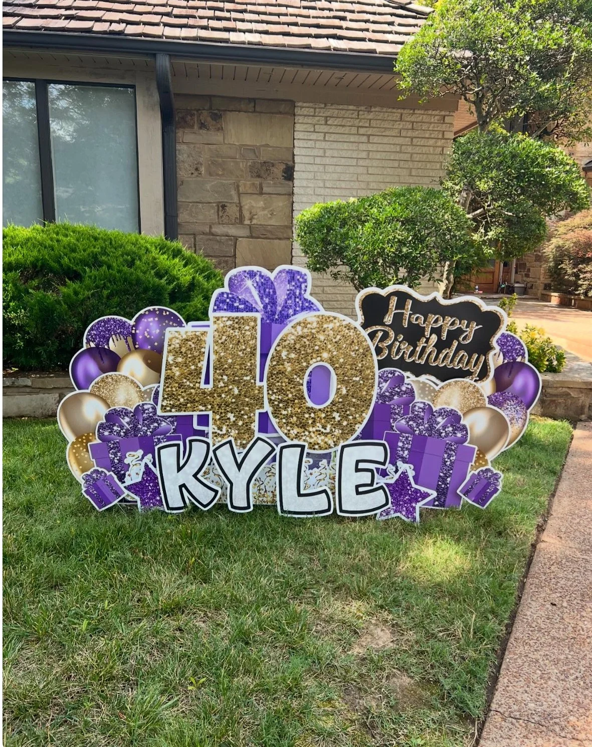 Outdoor birthday decoration with a sign reading "40 KYLE" in large gold glitter numbers, surrounded by purple and gold balloons, gift boxes, and a "Happy Birthday" message on a black background in a suburban yard.