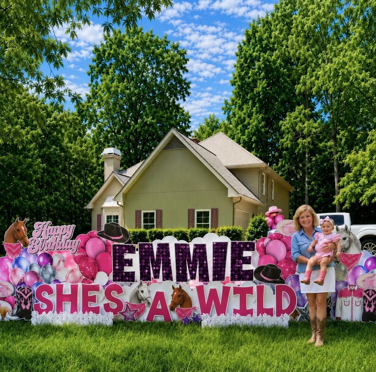 A woman and a young girl standing in front of a colorful birthday yard sign that reads "EMMIE SHE'S A WILD" with pink, purple, and blue balloons, hats, and animal decorations, in a residential backyard with a house, trees, and a blue sky in the backg