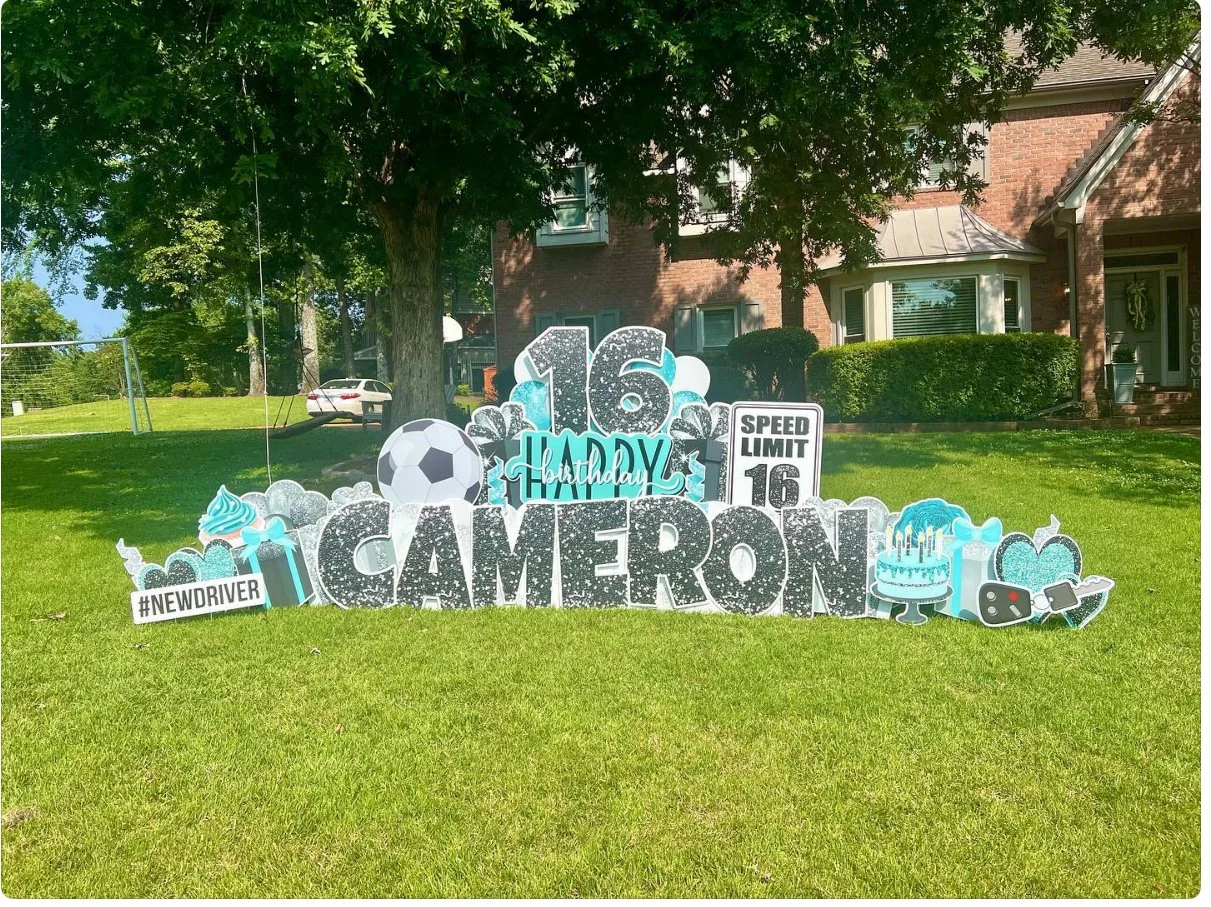 Birthday yard display with the name 'Cameron,' a large number 16, and soccer-themed decorations including a soccer ball, cupcake, and presents. There are also signs for speed limit 16, #NEWDRIVER, and celebration icons like cakes and cameras, on a gr