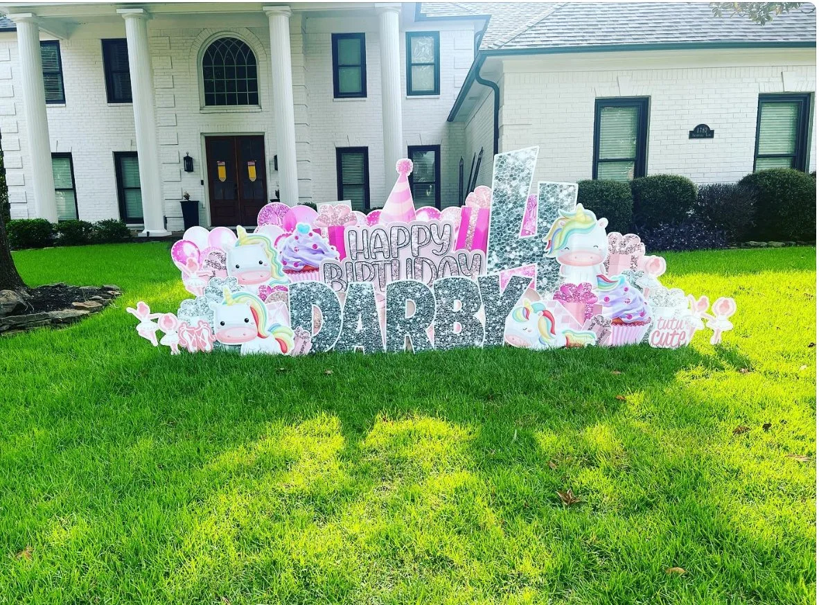 Pink and white birthday decoration on a lawn with unicorn and cupcake motifs, including the message "Happy Birthday Darby" in glittering letters, in front of a white house with columns.
