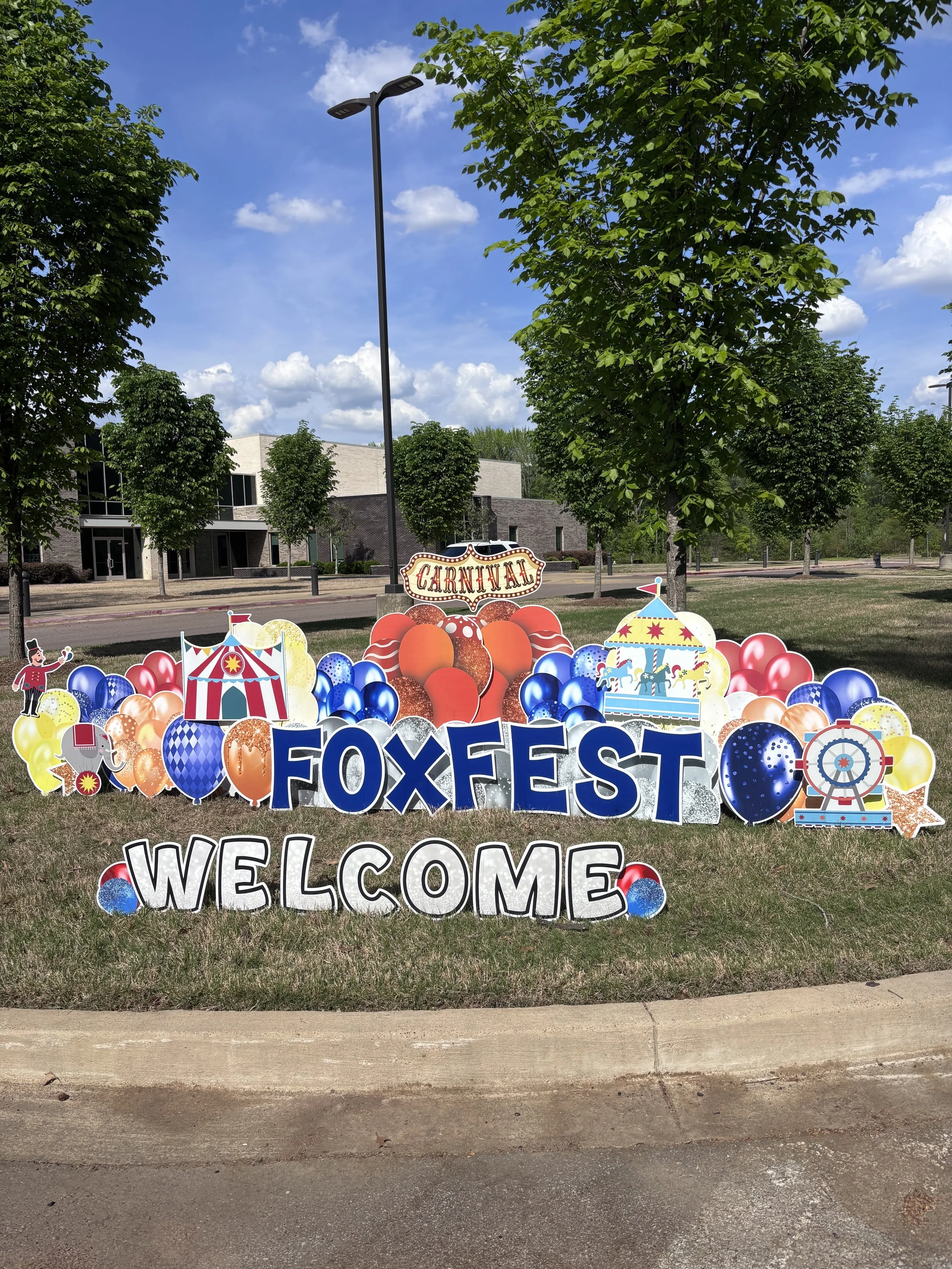 Decorative sign for a carnival and old west themed event at Foxtest, featuring colorful balloons, circus tents, a clown, and a rocking horse, with trees, buildings, and a blue sky in the background.