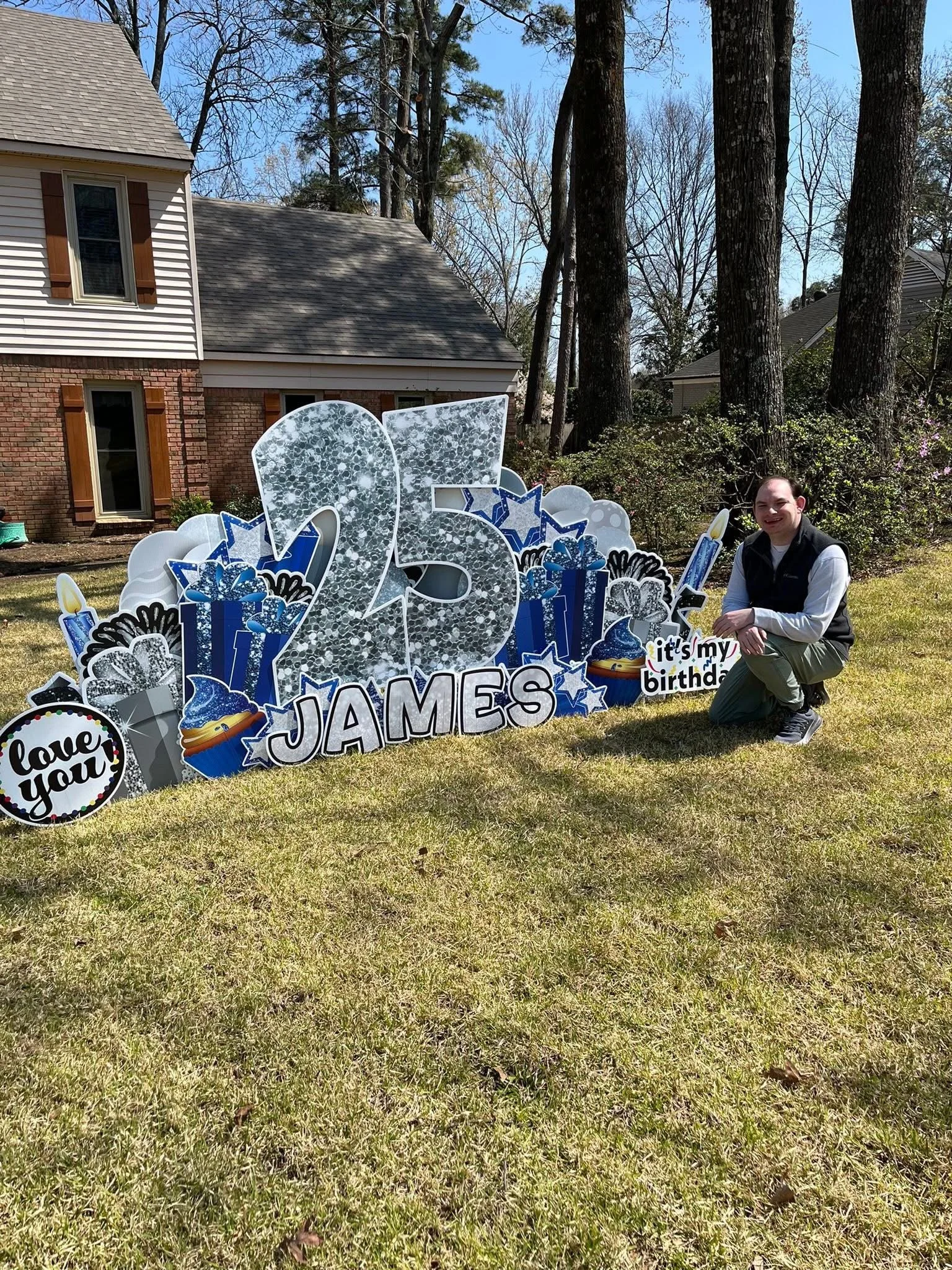 A man crouching next to large birthday decorations in a yard with grass, bare trees, and a house in the background. The decorations include the number 25 and themed birthday elements like presents, cupcakes, and candles.