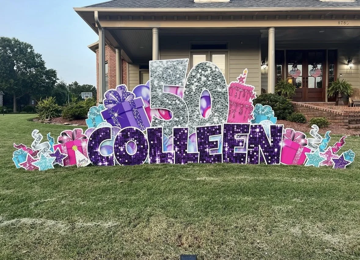 Decorative outdoor display celebrating Colleen's 50th birthday with large silver, purple, and pink numbers, gifts, stars, and birthday cake cutouts in front of a house with a porch.