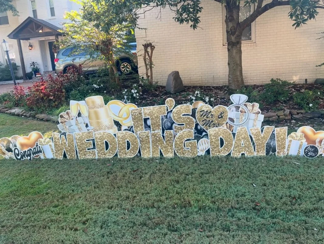 Decorative sign on lawn with the words 'It's Wedding Day!' and celebratory symbols, including champagne glasses, a top hat, rings, and hearts.