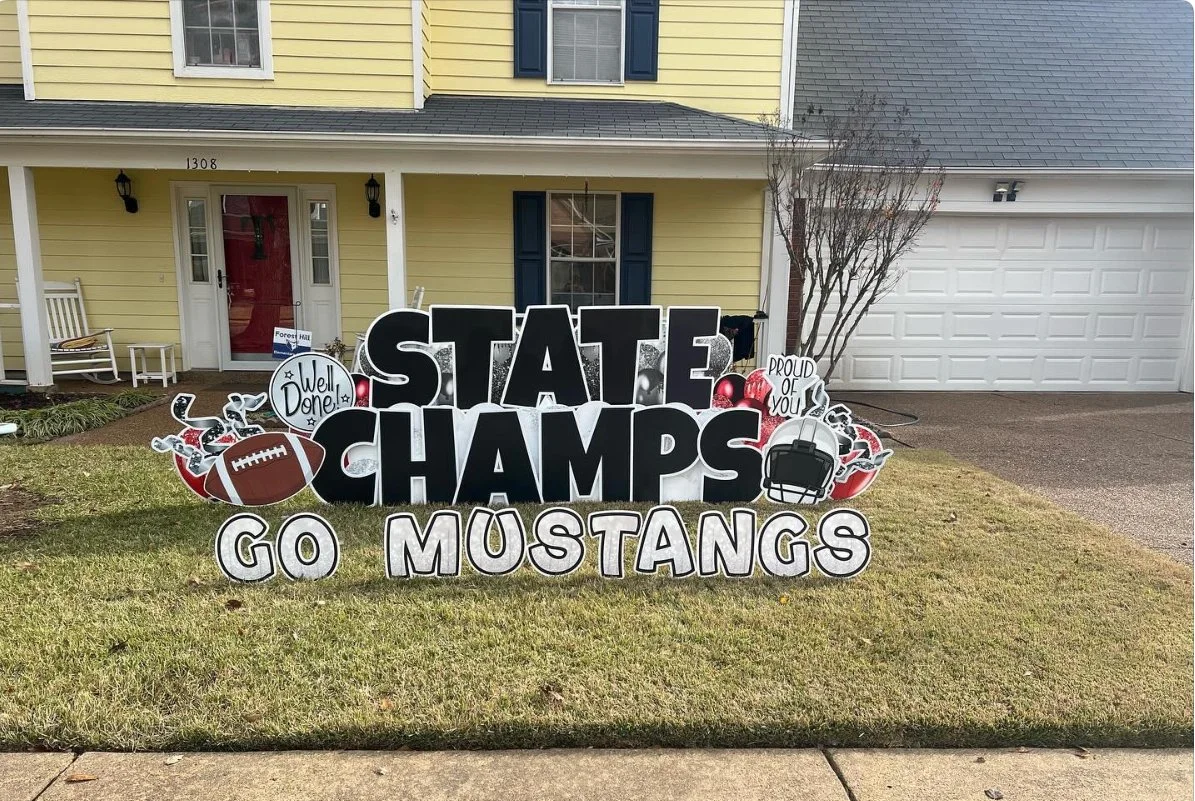 Decorative yard sign supporting a team, with phrases like 'State Champs' and 'Go Mustangs,' featuring football and football helmet graphics in front of a yellow house with a white garage door.