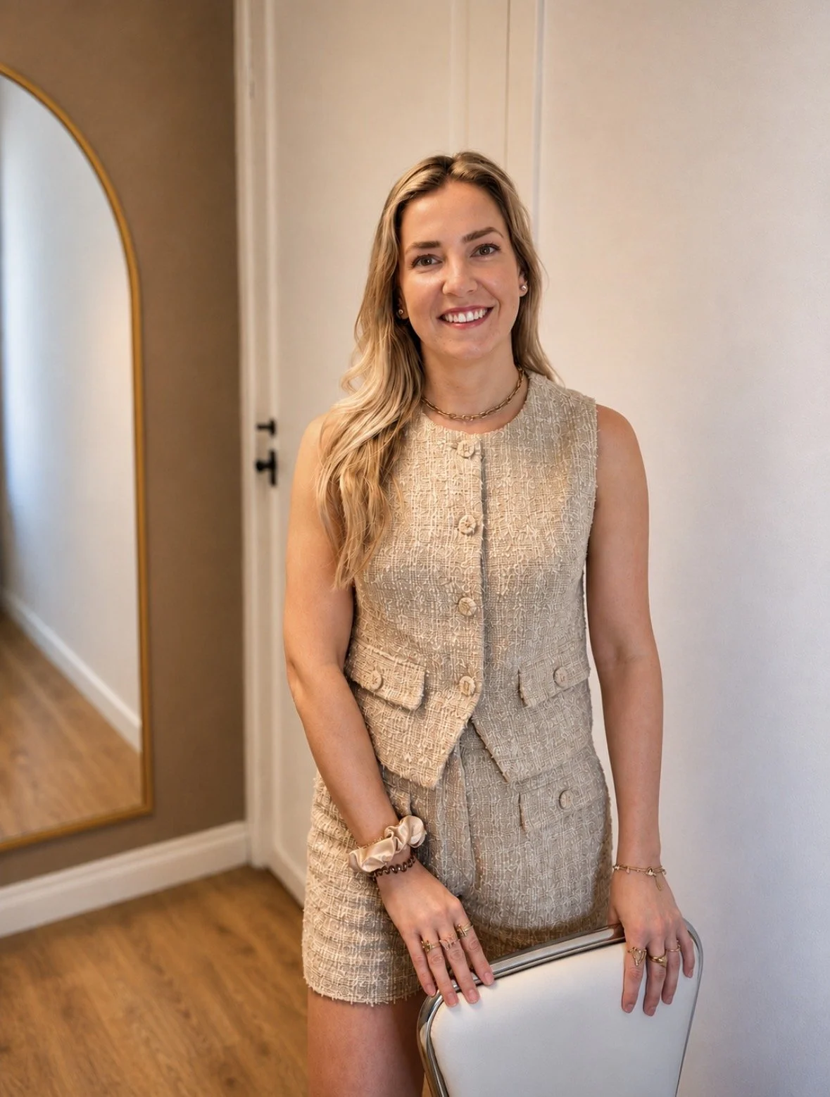 Een lachende jonge vrouw met licht bruin haar in een beige outfit, binnen een huis met houten vloer en een spiegeltje op de muur.