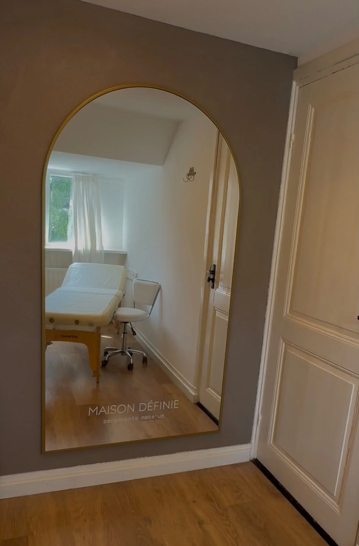 Een kamer met een ziekenhuisbed, een witte bureaustoel en een raam met witte gordijnen, zichtbaar door een grote spiegel.