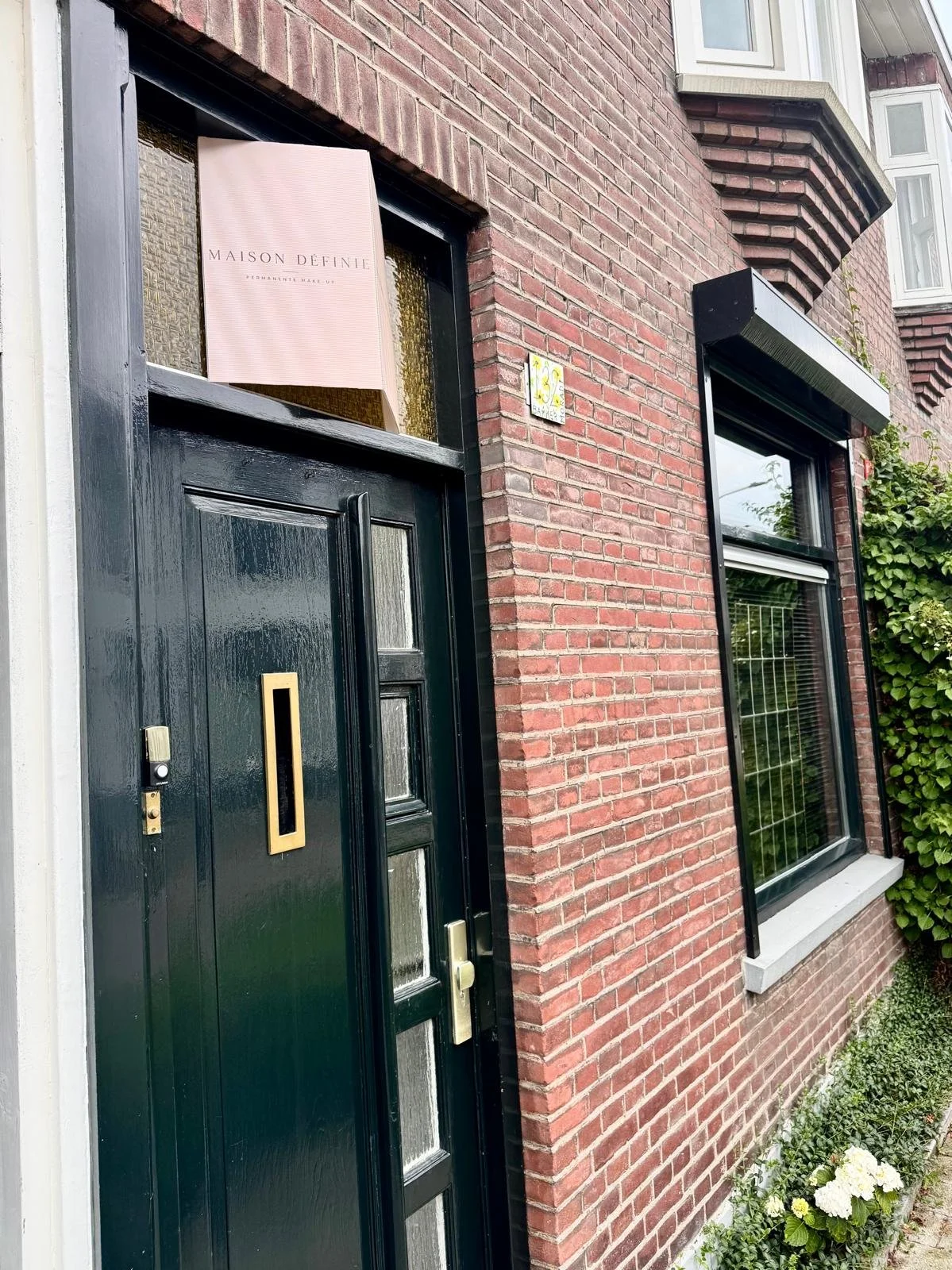Een rode bakstenen woning met een zwarte voordeur en een raam met een andere raamdecoratie. Boven het raam hangt een bord met de tekst 'MAISON DÉFINIE'.