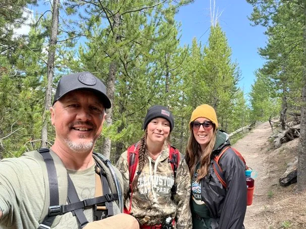 Three people taking a selfie outdoors on a hiking trail surrounded by green trees, wearing outdoor gear and backpacks, smiling.