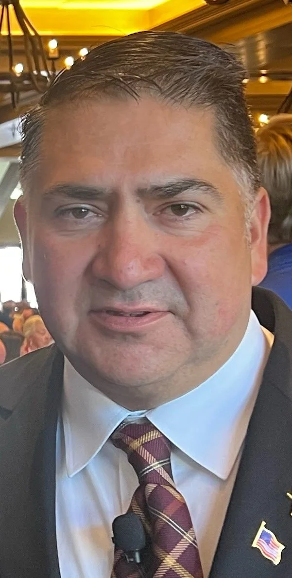 A man with dark hair, wearing a white shirt, a patterned maroon and yellow tie, and a dark suit jacket, standing in an indoor setting with warm lighting and people in the background.