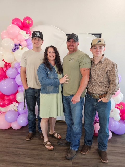 A family of four posing in front of a balloon decoration with pink, purple, and white balloons and flowers, on a wooden floor, with a white backdrop.
