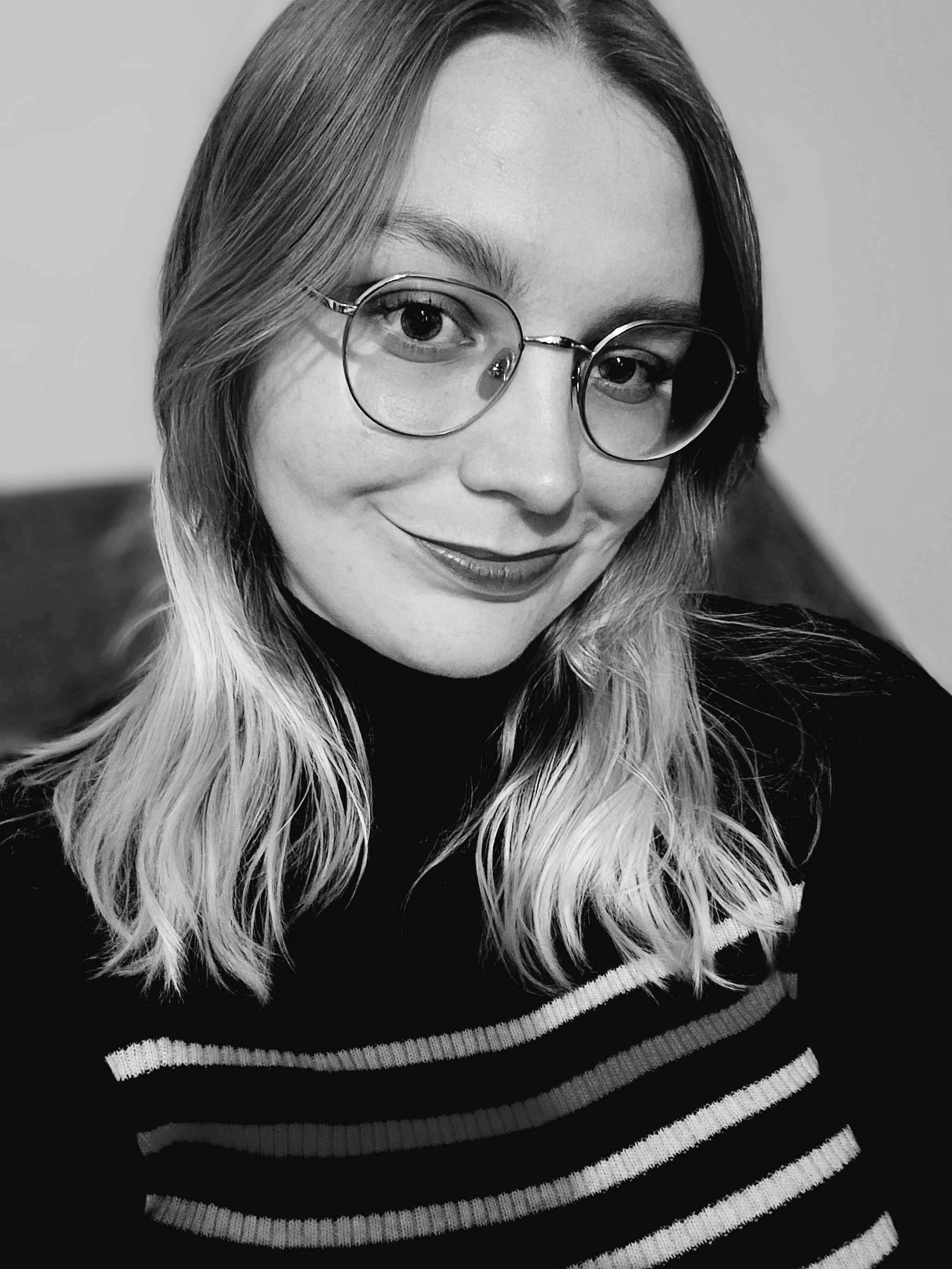 A close-up black and white photo of a smiling person wearing round glasses and a striped sweater. They have long, wavy hair and are looking at the camera with a cheerful expression.