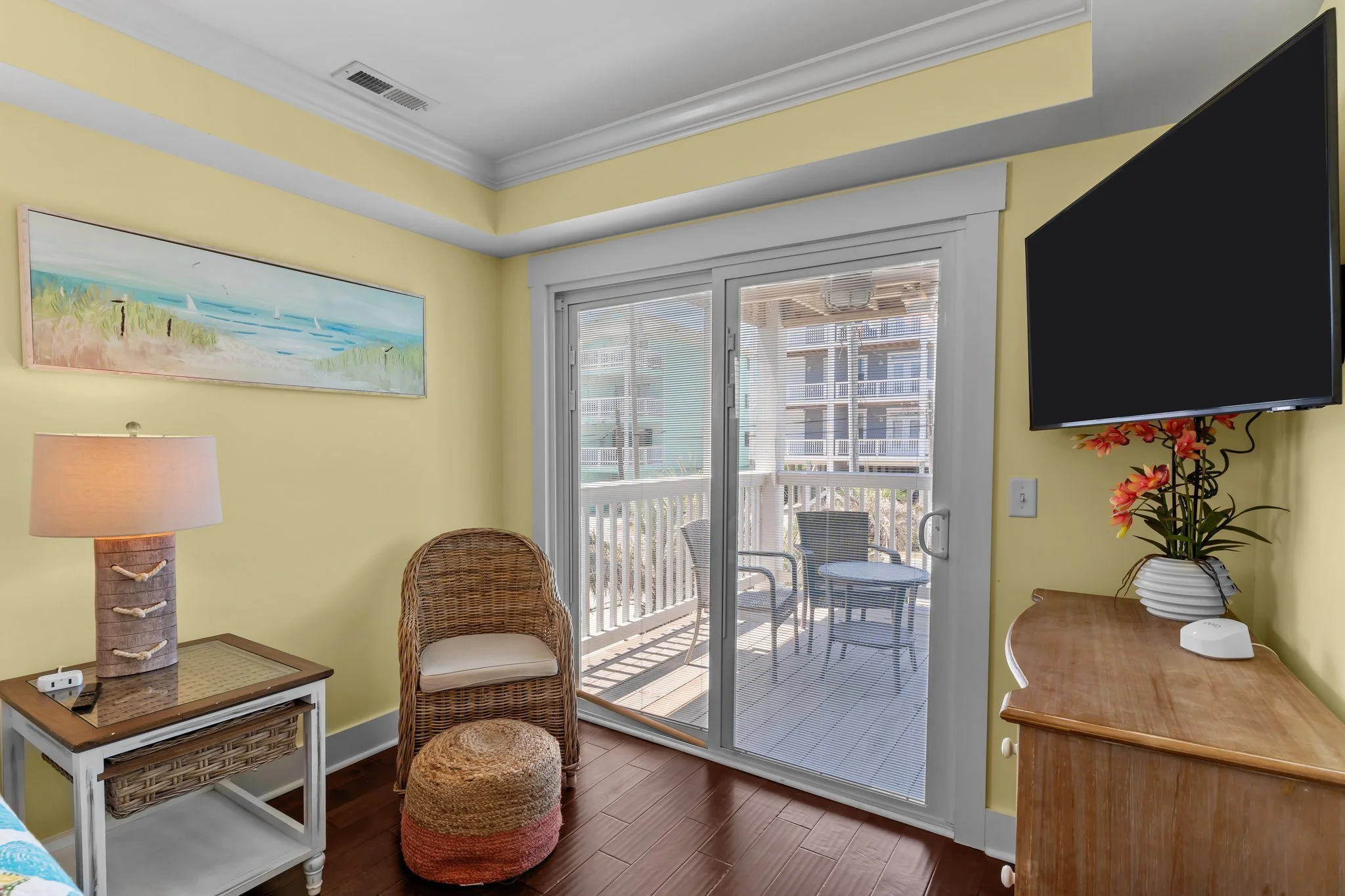 A cozy corner of a living room with pastel yellow walls, a wicker chair, a small white side table with a woven basket-like lamp, a round woven ottoman, a sliding glass door leading to a balcony, a vibrant floral plant in a white vase, a black flat-sc
