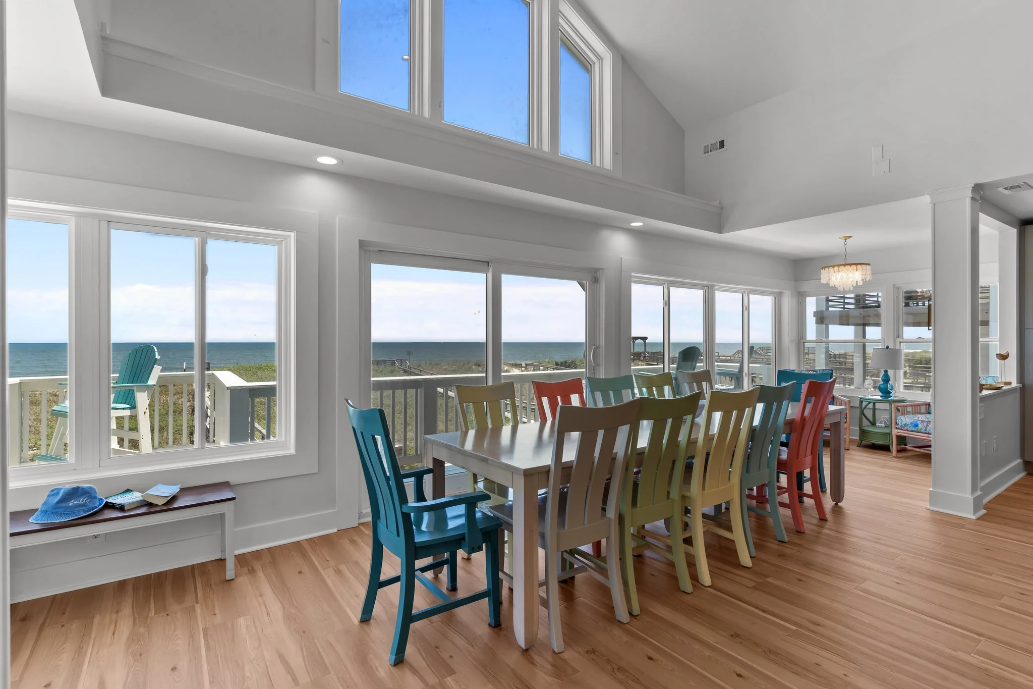 Bright dining area with multicolored chairs, white table, large windows facing ocean, and a view of the beach and ocean outside.