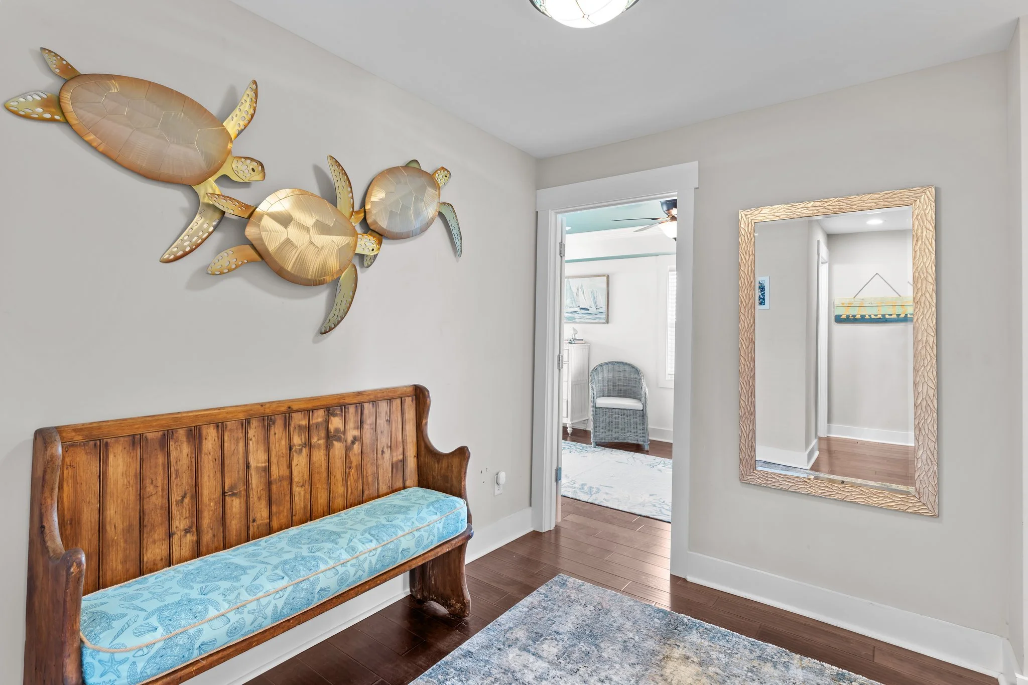 Interior of a room featuring a wooden bench with a light blue cushion, white walls, a mirror with a textured beige frame, and a metal wall art piece of three sea turtles.