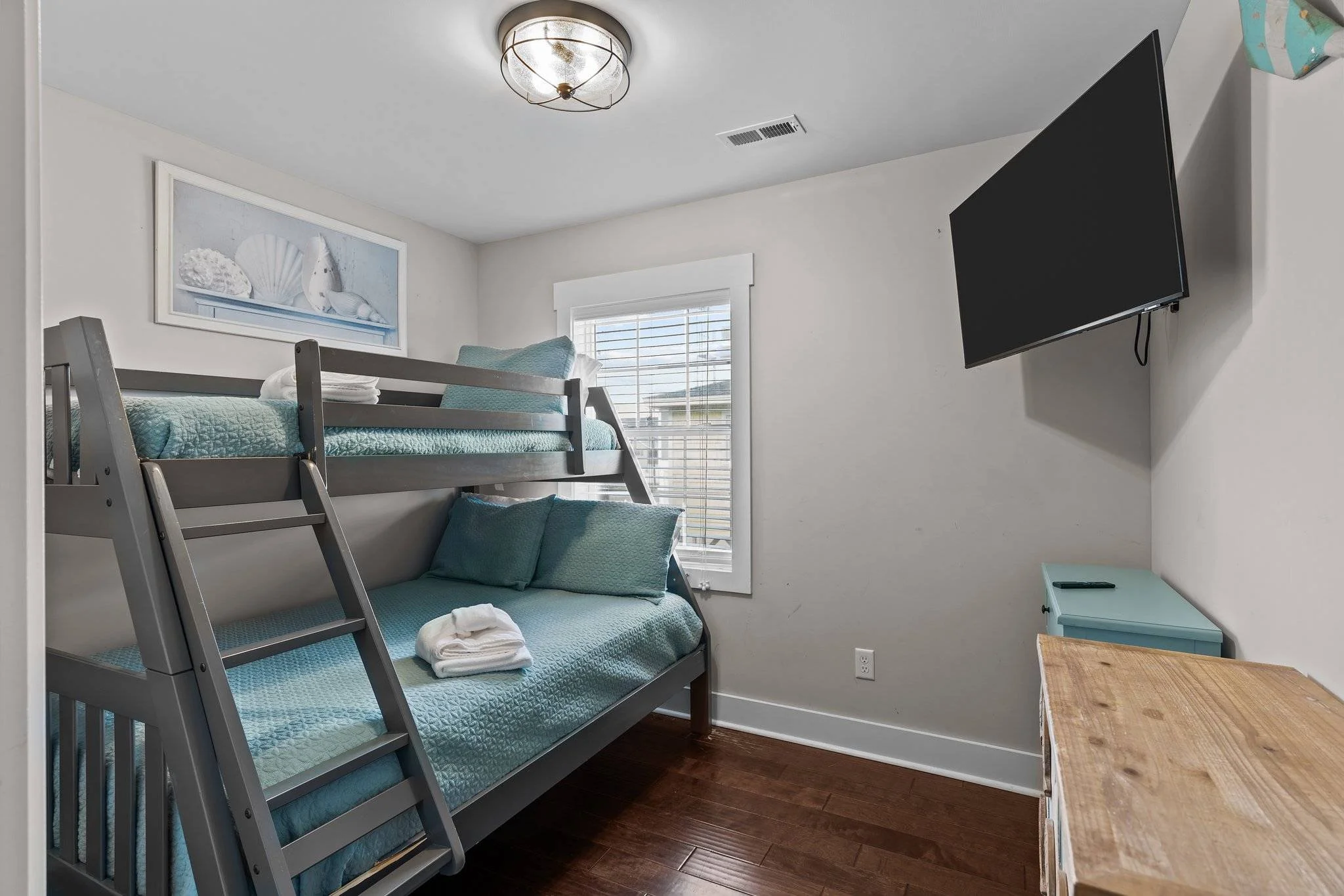 A bedroom with a bunk bed and a wall-mounted flat-screen TV. The room has a light-colored wall, a window with blinds, a ceiling light, and a small teal nightstand. The top bunk has a quilted teal mattress with pillows, while the bottom bunk has pillo