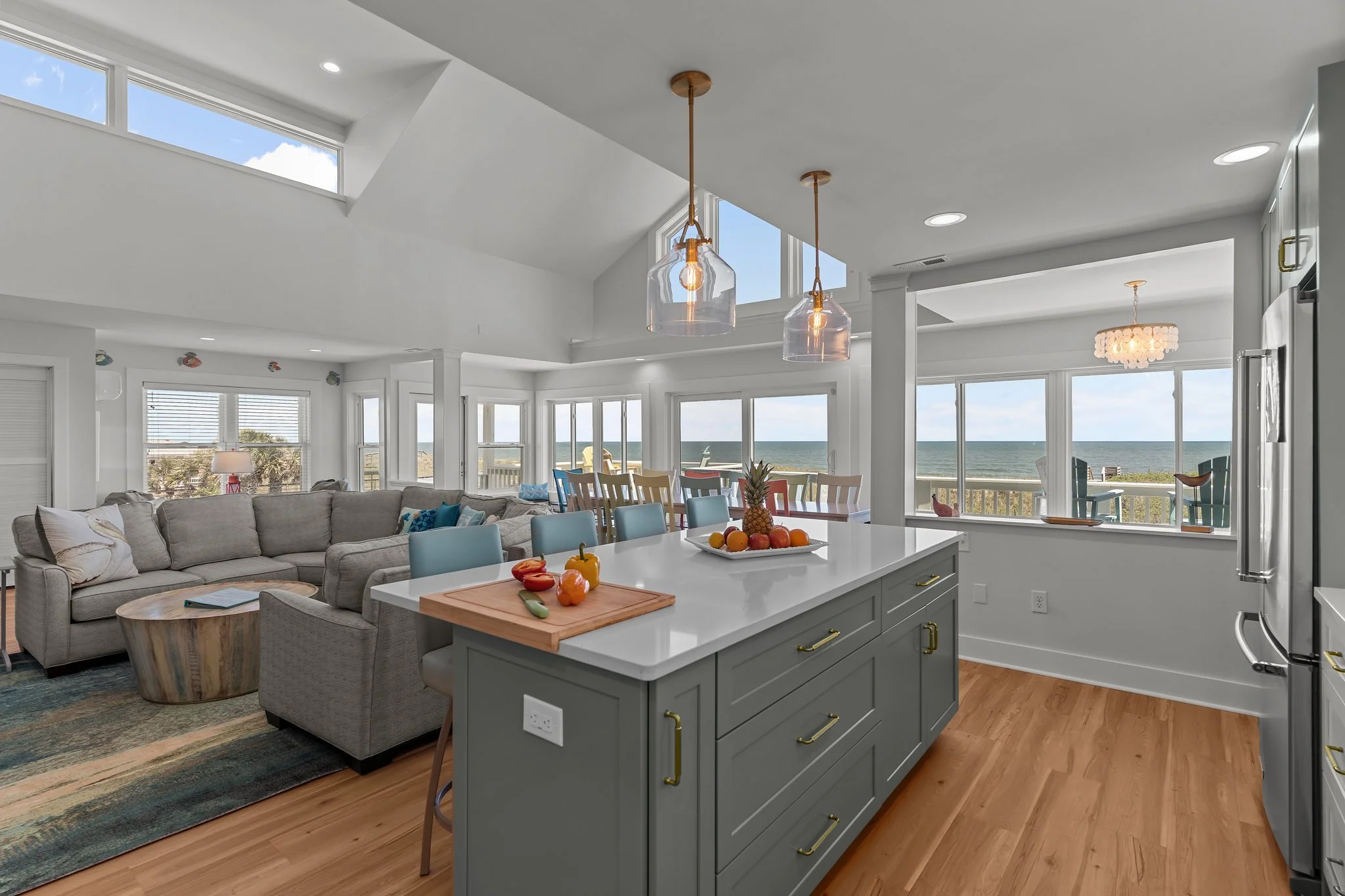 Open-concept living room and kitchen with ocean view, white walls, modern lighting, and a large kitchen island with fruits and vegetables on top.