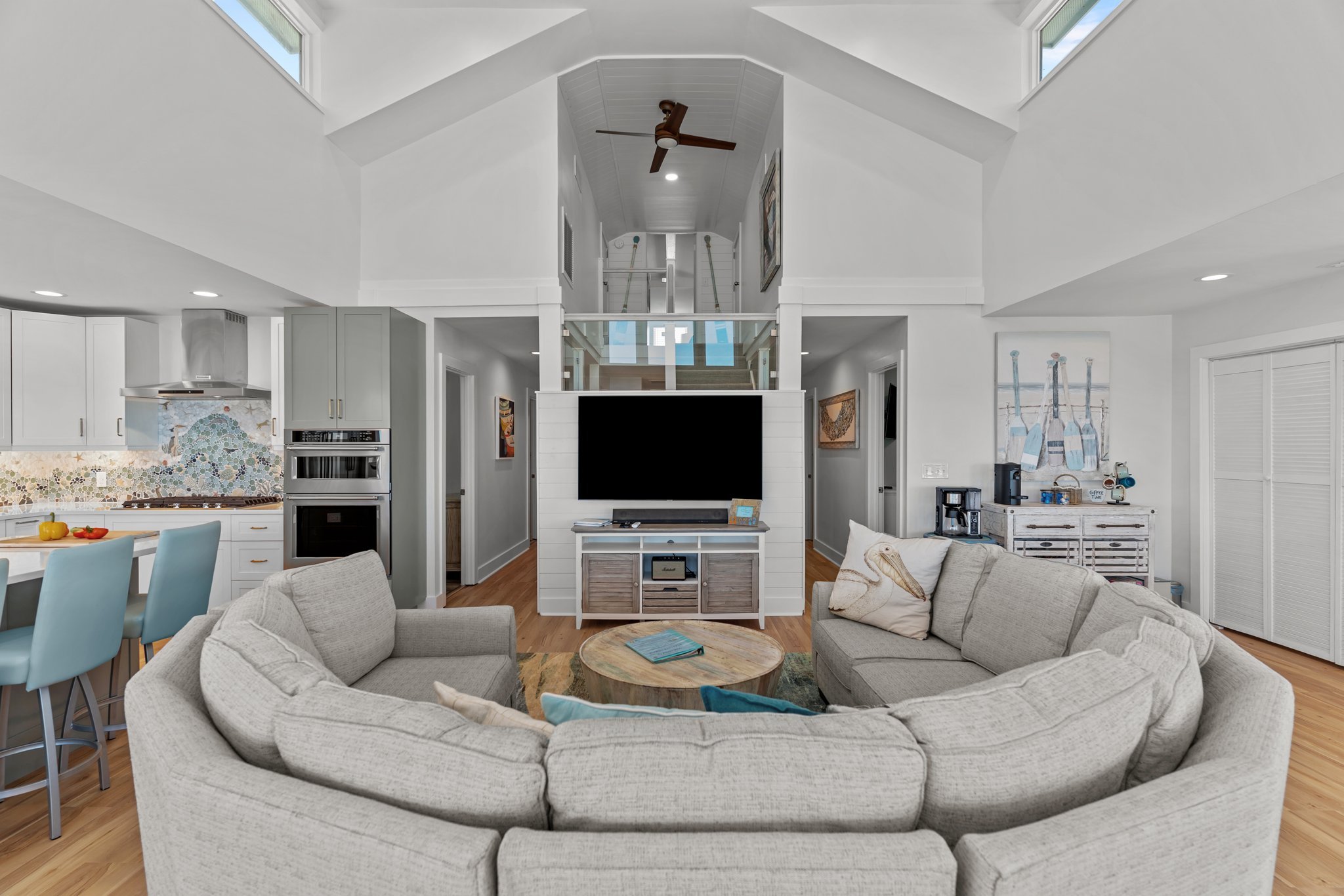Living room with beige sectional sofa, coffee table, wall-mounted TV, kitchen with white cabinets, and high vaulted ceiling with skylights and a ceiling fan.