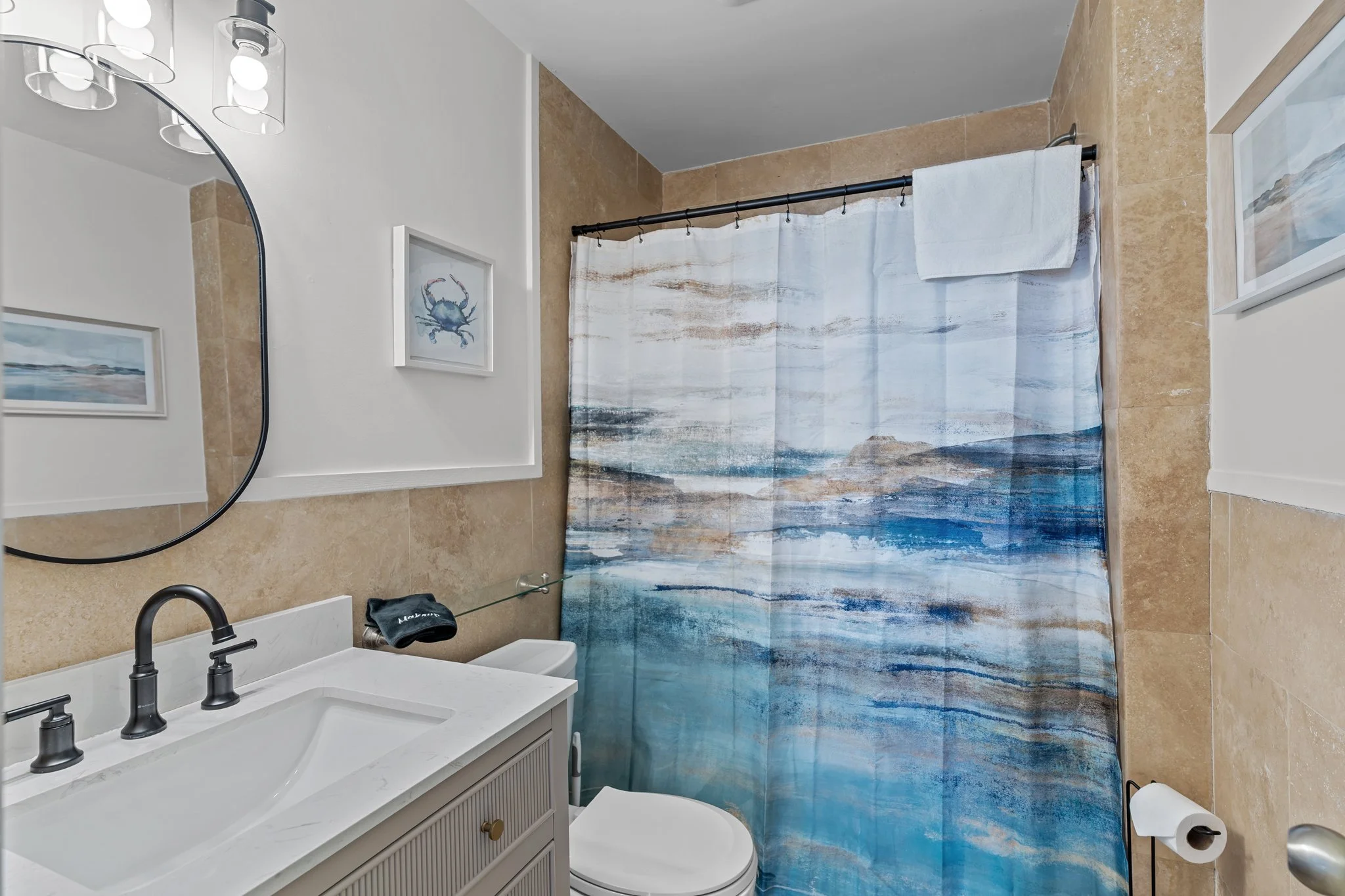 A bathroom with a white vanity, black faucet, and a large round mirror. The shower has a curtain with a seascape design, and there are framed ocean-themed pictures on the wall. The walls are tiled in beige, and a towel hangs on a rod near the shower.