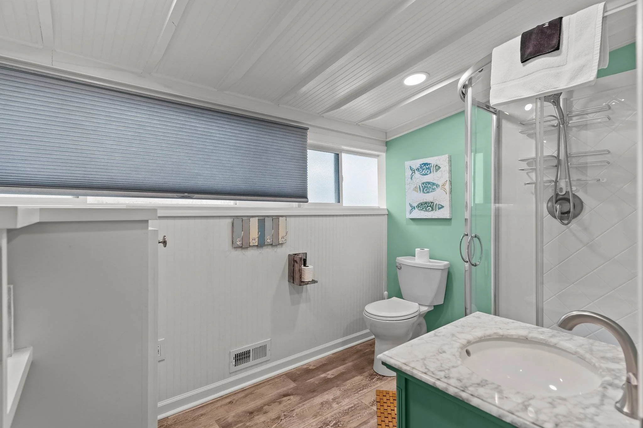 A clean bathroom with white beadboard walls and ceiling, green accent wall, a white toilet, a marble countertop sink, a shower with glass door, and a window with a blue-gray horizontal blind.